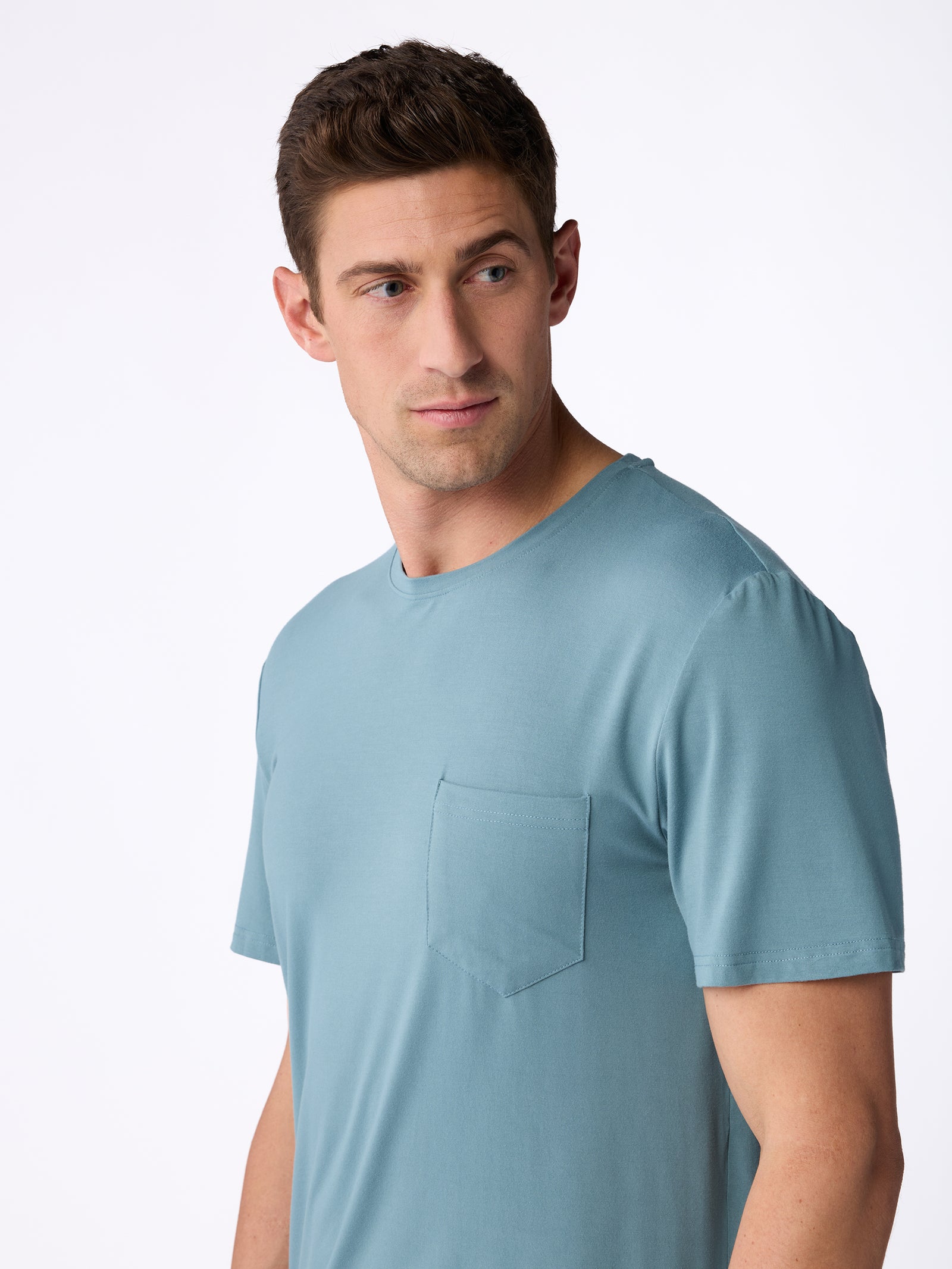 A man with short brown hair is wearing a Men's Stretch-Knit Bamboo Lounge Tee in light blue from Cozy Earth, featuring a chest pocket. He is looking off to the side against a plain white background. 