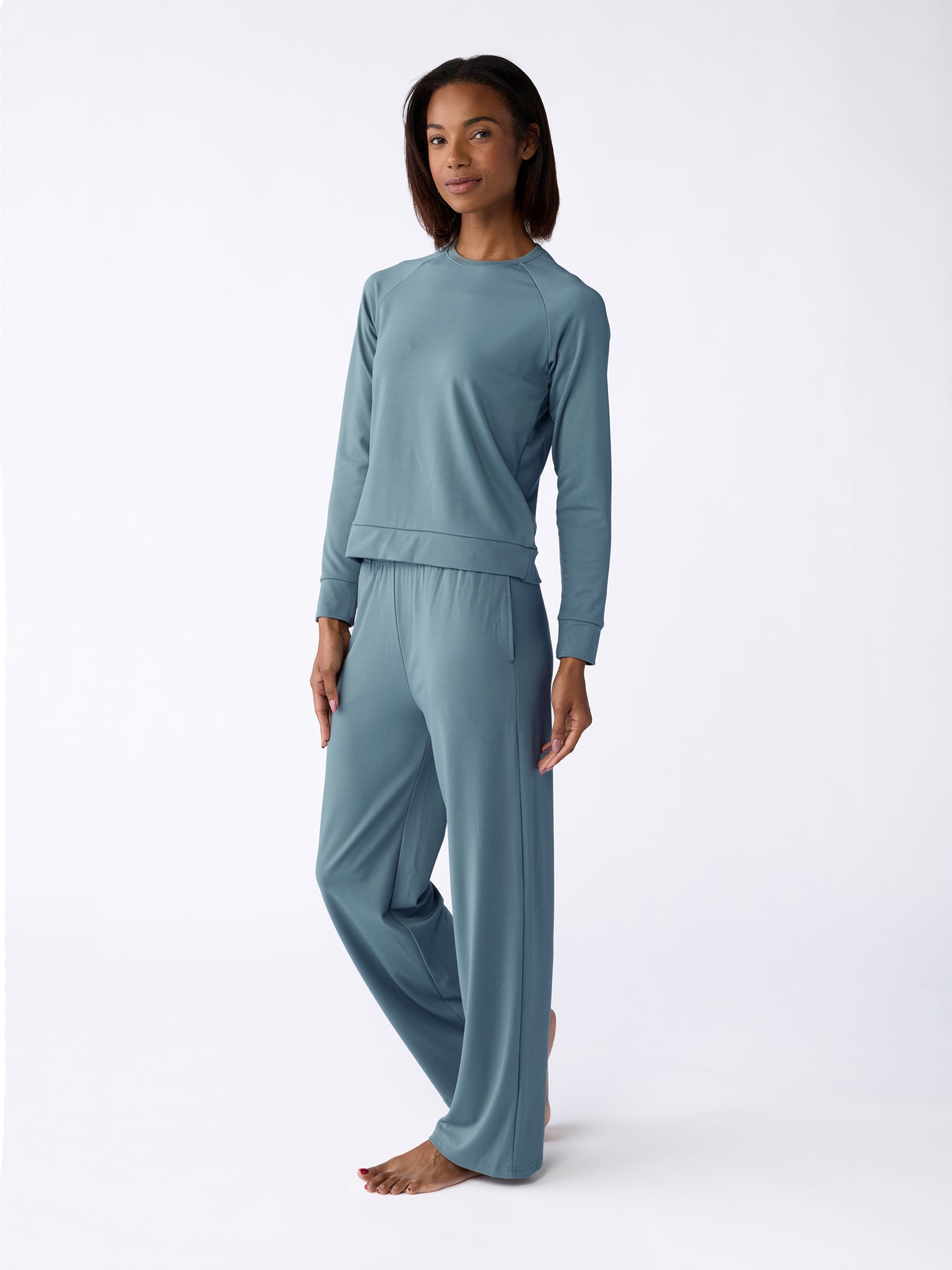 A woman wearing the Cozy Earth Women's Ultra-Soft Bamboo Wide Leg Pull On Pant & Pullover Crew Set in light teal stands barefoot against a plain white background, exhibiting a relaxed expression and neutral posture. |Color:Oceanside