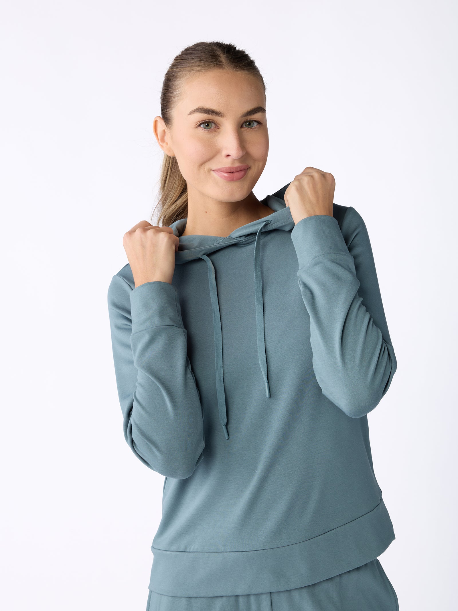 A person wearing the Women's Ultra-Soft Bamboo Hoodie by Cozy Earth stands against a white background, holding the hood strings with both hands. They have long hair pulled back and a slight smile on their face. 