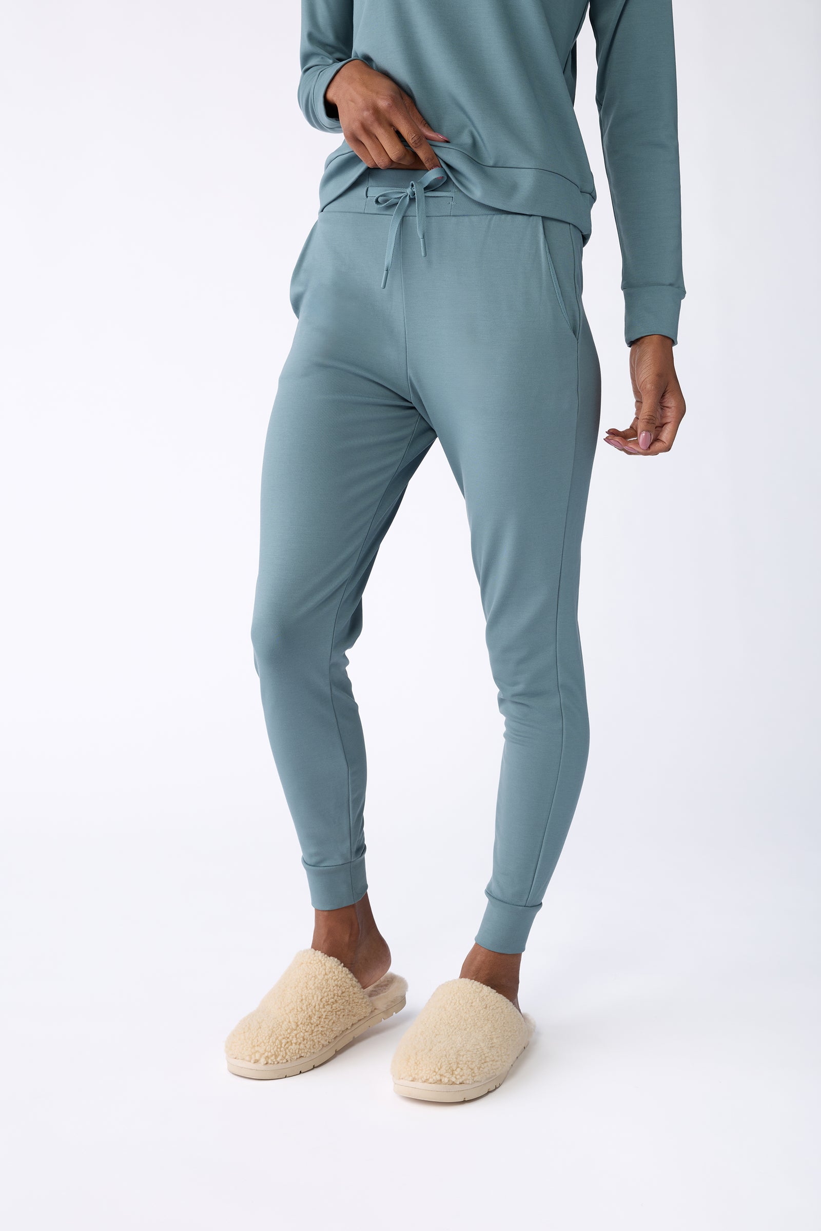 A person is seen wearing Cozy Earth's Women's Bamboo Jogger Pant in teal and a matching sweatshirt, standing with one hand in their pocket. They also have on a pair of beige fuzzy slippers, all set against a plain white background. The image highlights the lower half of the body. 