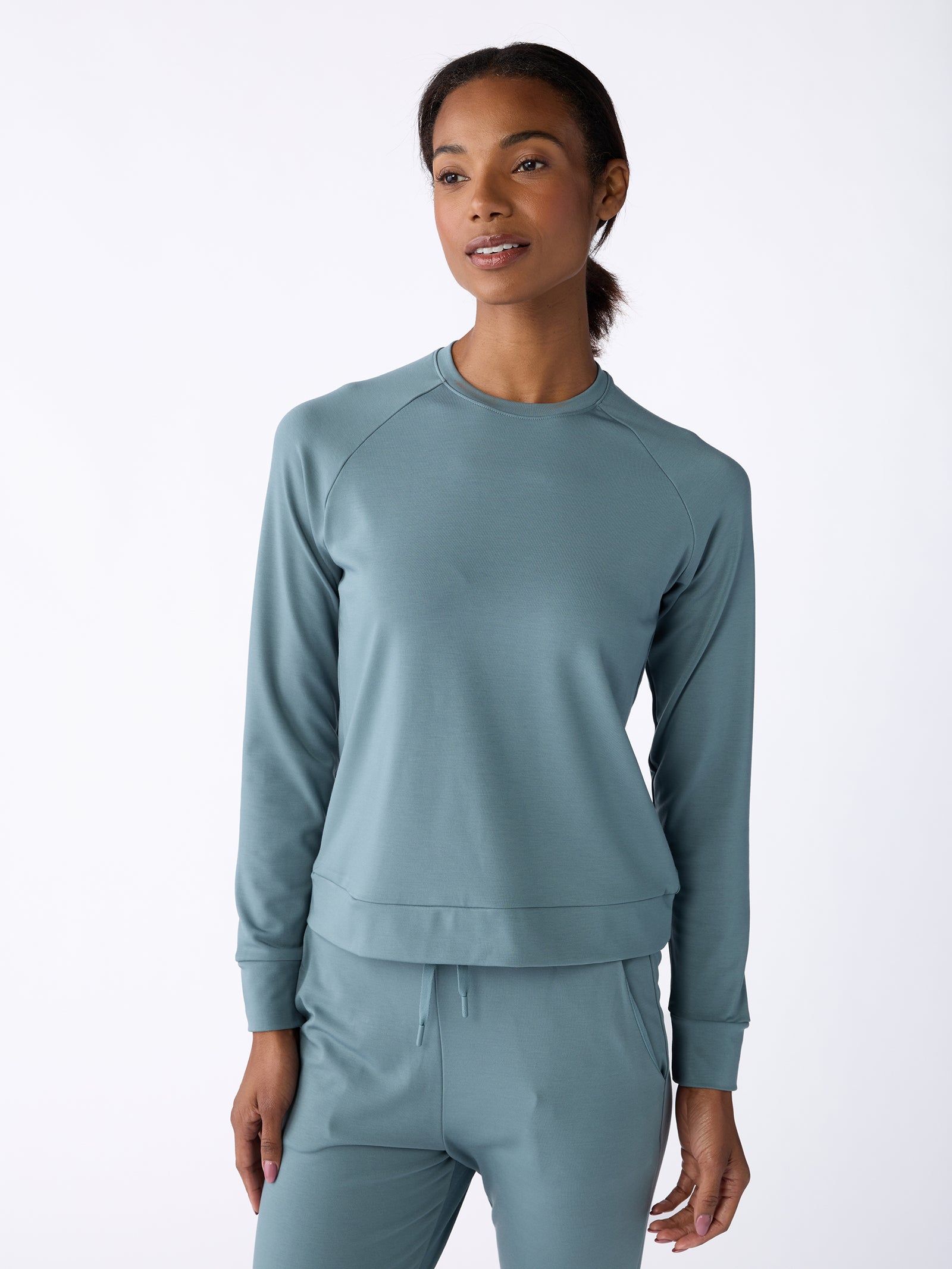 A person wearing a light blue Women's Ultra-Soft Bamboo Pullover Crew by Cozy Earth and matching pants stands against a plain white background. The outfit appears comfortable and casual. 