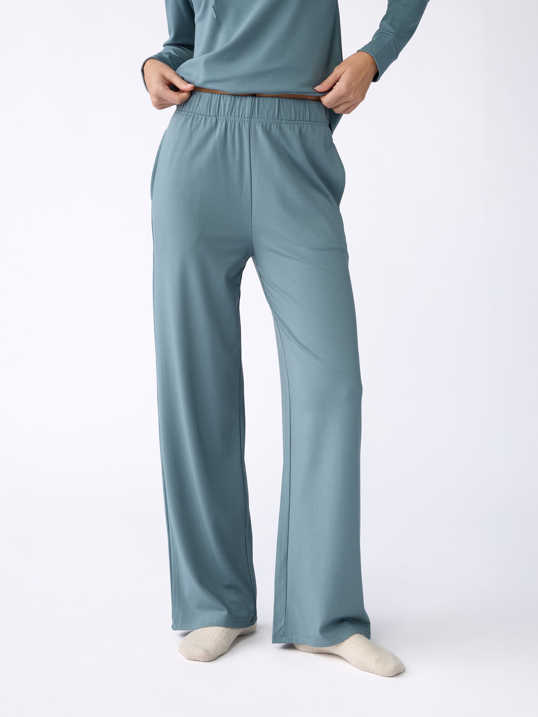 A person is standing against a plain white background, wearing the Women's Ultra-Soft Bamboo Wide Leg Pull On Pant in light blue from Cozy Earth, with one hand in their pocket. They are also wearing cream-colored socks and a matching top. |Color:Oceanside