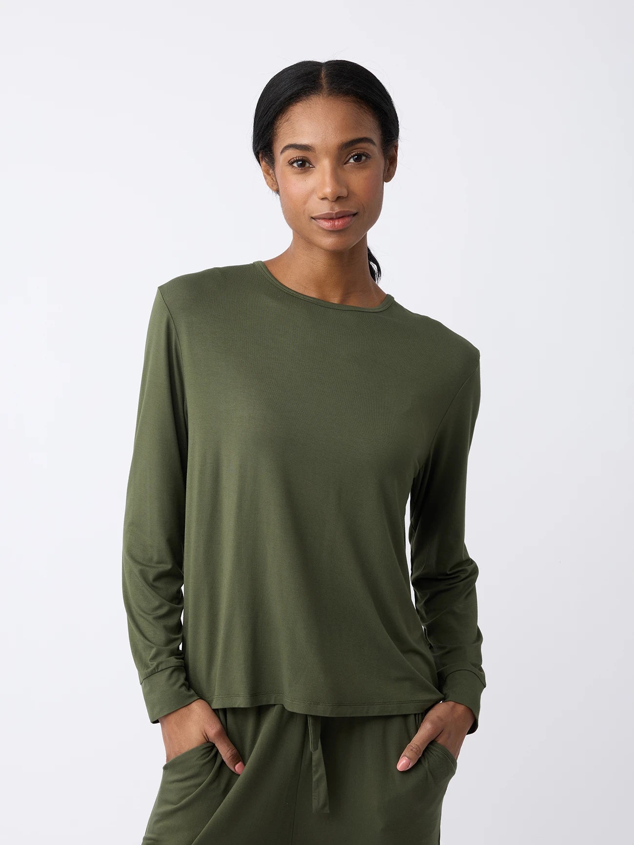 A woman stands against a white background wearing the Women's Stretch Knit Bamboo Long Sleeve Lounge Tee by Cozy Earth, paired with matching pants. They have long, dark hair pulled back and smile softly with hands in their pockets. 