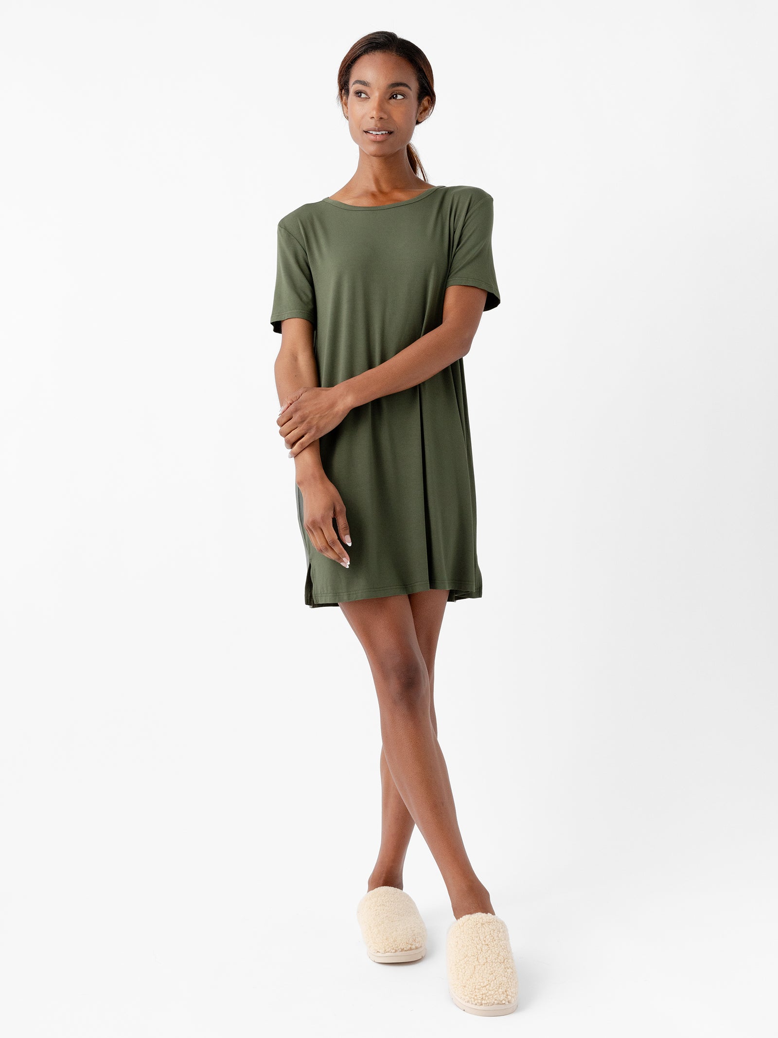 A woman stands against a white background, wearing the Women's Bamboo Stretch Knit Sleep Dress from Cozy Earth in green, paired with beige fuzzy slippers. She has her arms gently crossed and is looking forward with a neutral expression. 