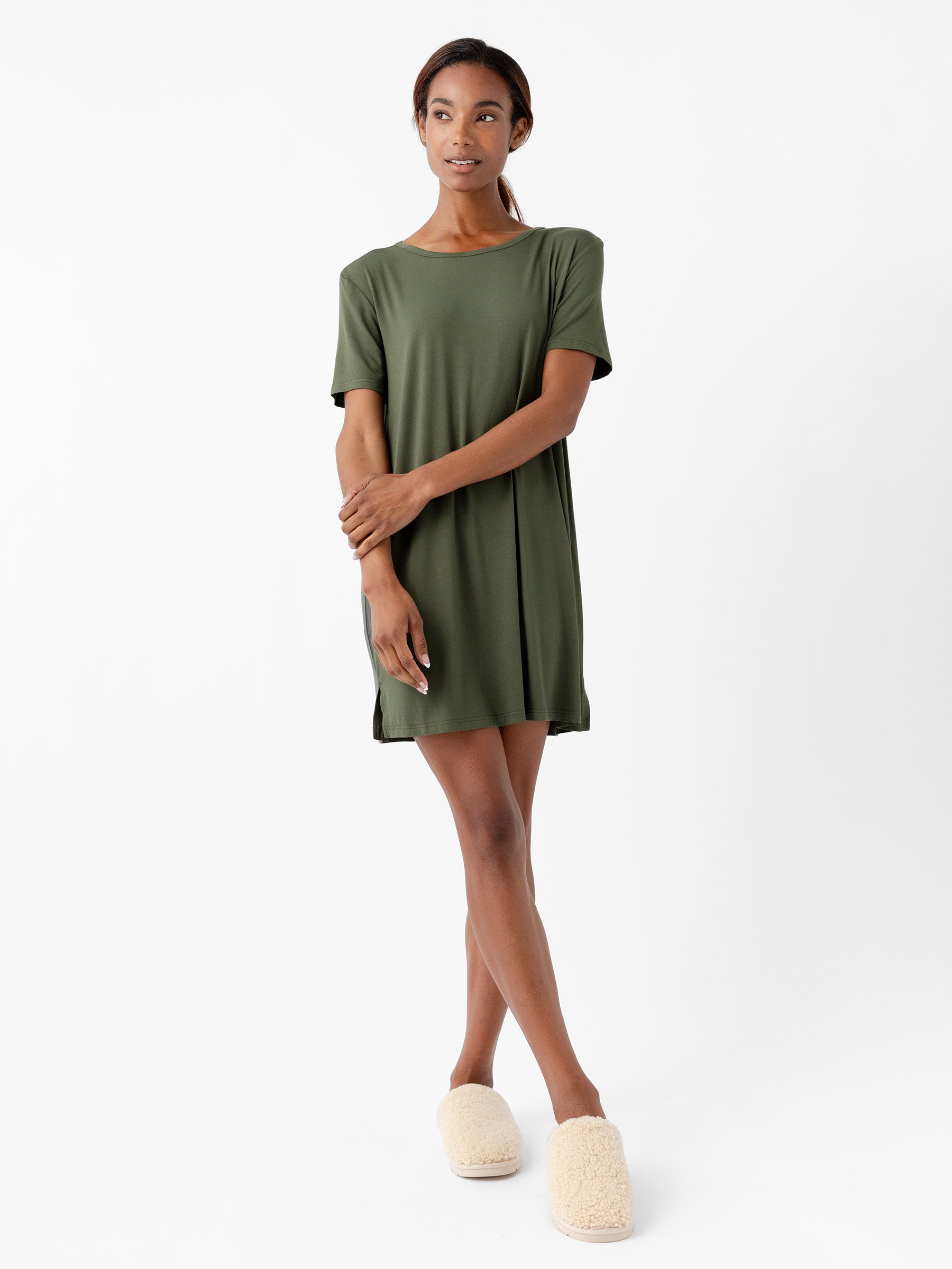 A woman stands against a white background, wearing the Women's Bamboo Stretch Knit Sleep Dress from Cozy Earth in green, paired with beige fuzzy slippers. She has her arms gently crossed and is looking forward with a neutral expression. |Color:Olive