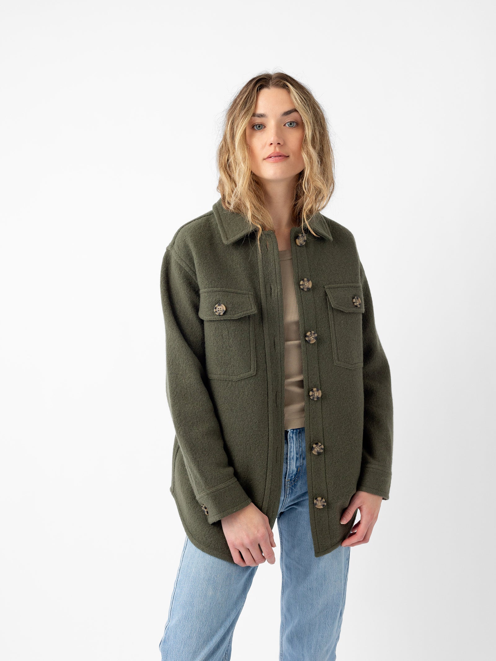 A person with wavy, shoulder-length hair wears the Cozy Earth Women's Boucle Shacket in olive green, characterized by its large front pockets. They pair it with a beige top and light blue jeans while standing against a plain white background, looking just past the camera with a neutral expression. 