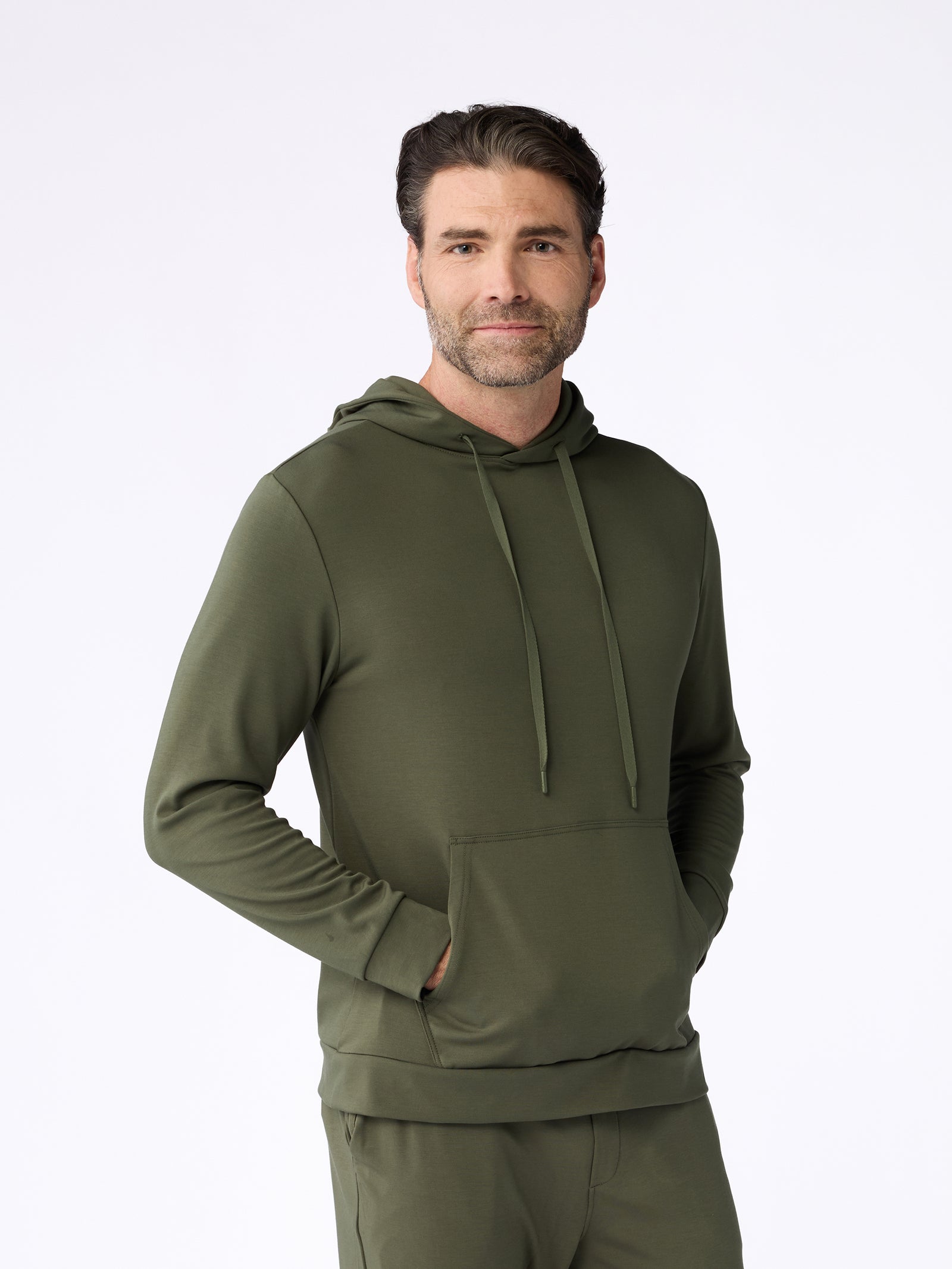 A man with short, dark hair wearing a Men's Ultra-Soft Bamboo Hoodie by Cozy Earth stands against a plain white background. He has his hand in the front pocket and is smiling slightly. 