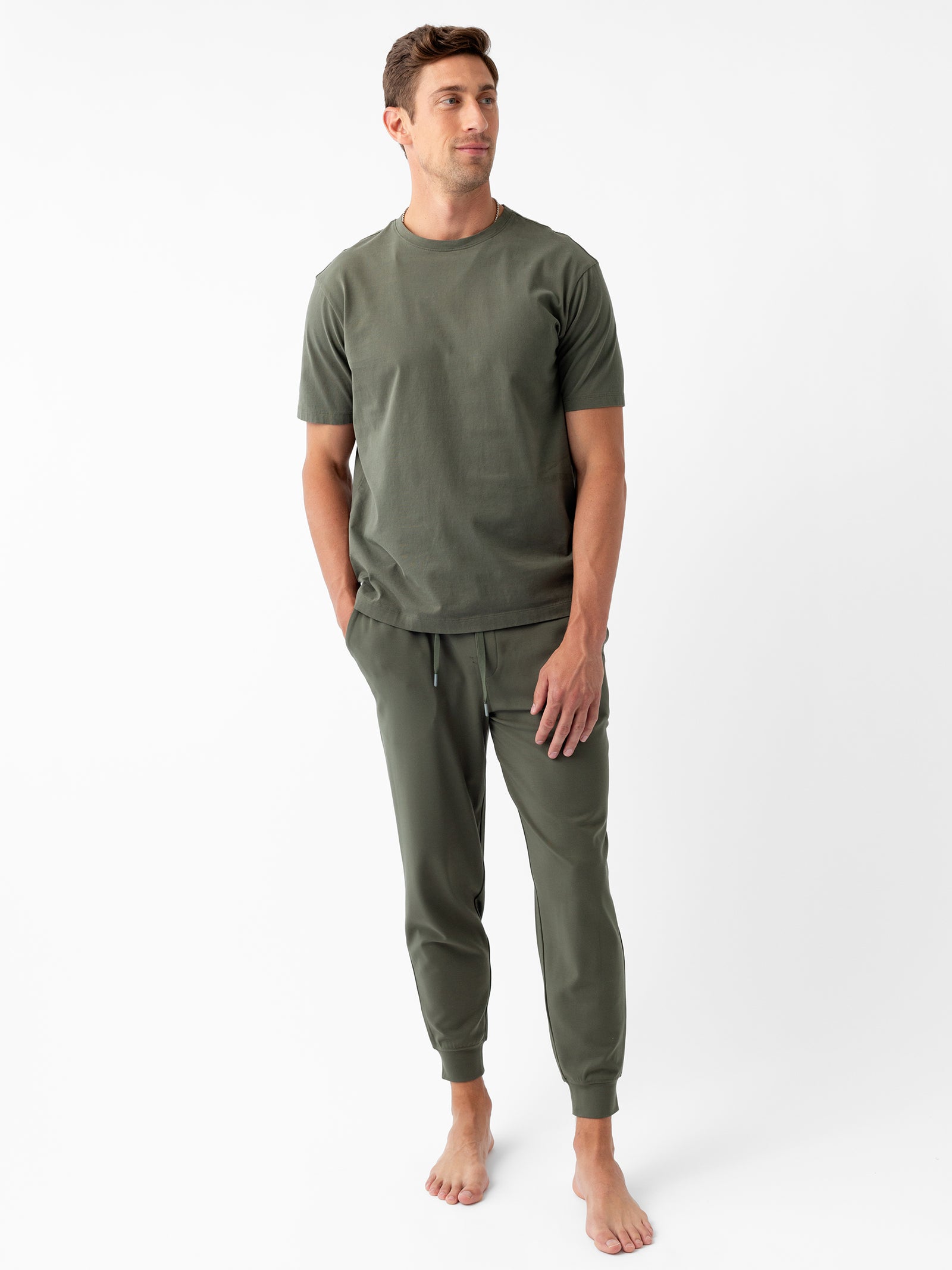 A person stands barefoot against a plain white background. They are wearing an olive-green, short-sleeve, crew-neck t-shirt paired with Cozy Earth's Men's Ultra-Soft Bamboo Jogger Pant. Their hands are in their pockets, and they are looking slightly to the side while smiling. 