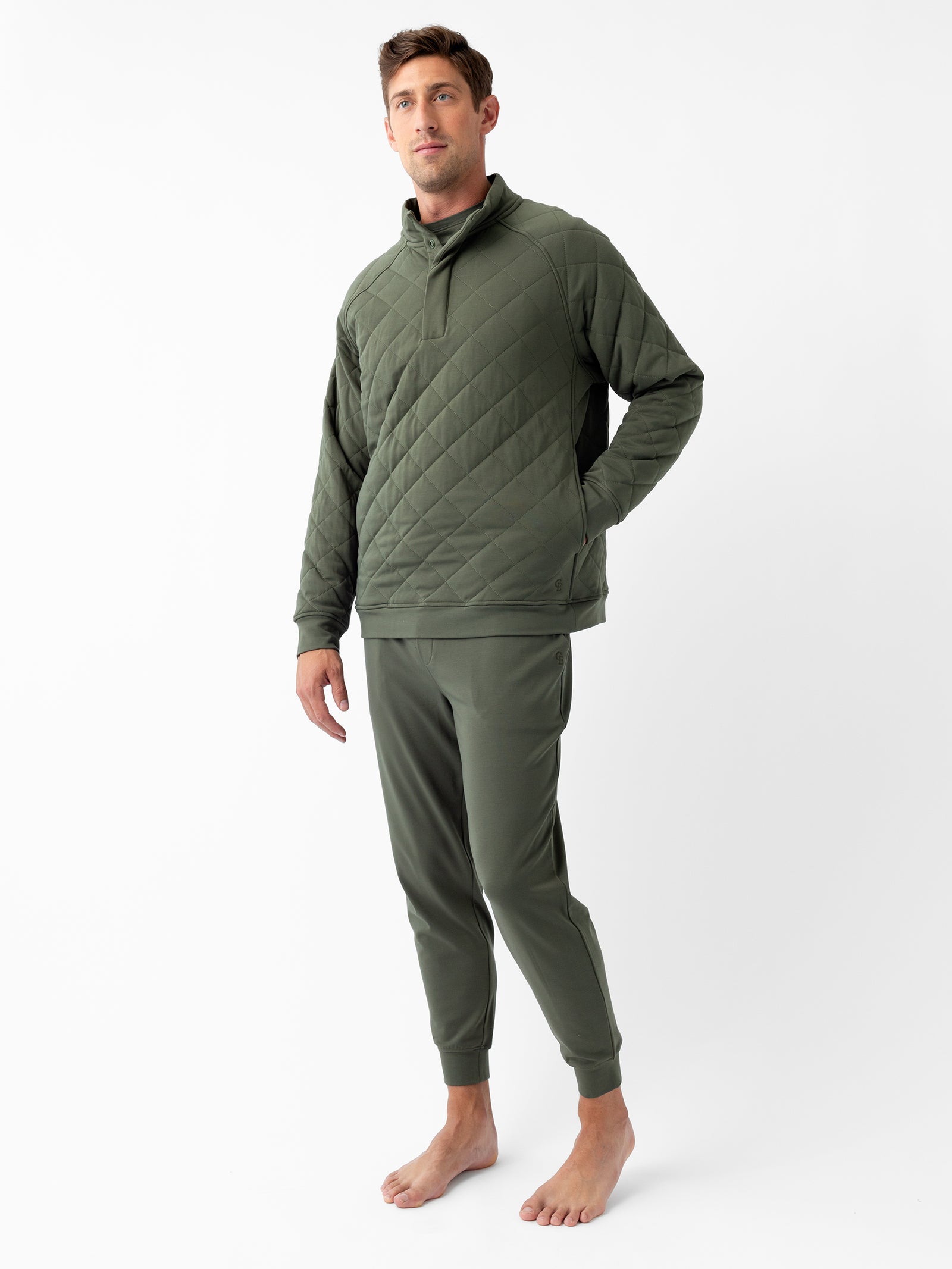 A man stands barefoot against a white background, wearing the Men's Ultra-Soft Bamboo Quilted Snap Pullover and matching dark green joggers from Cozy Earth. He poses with one hand relaxed by his side and the other in his pocket, looking off to the side. 