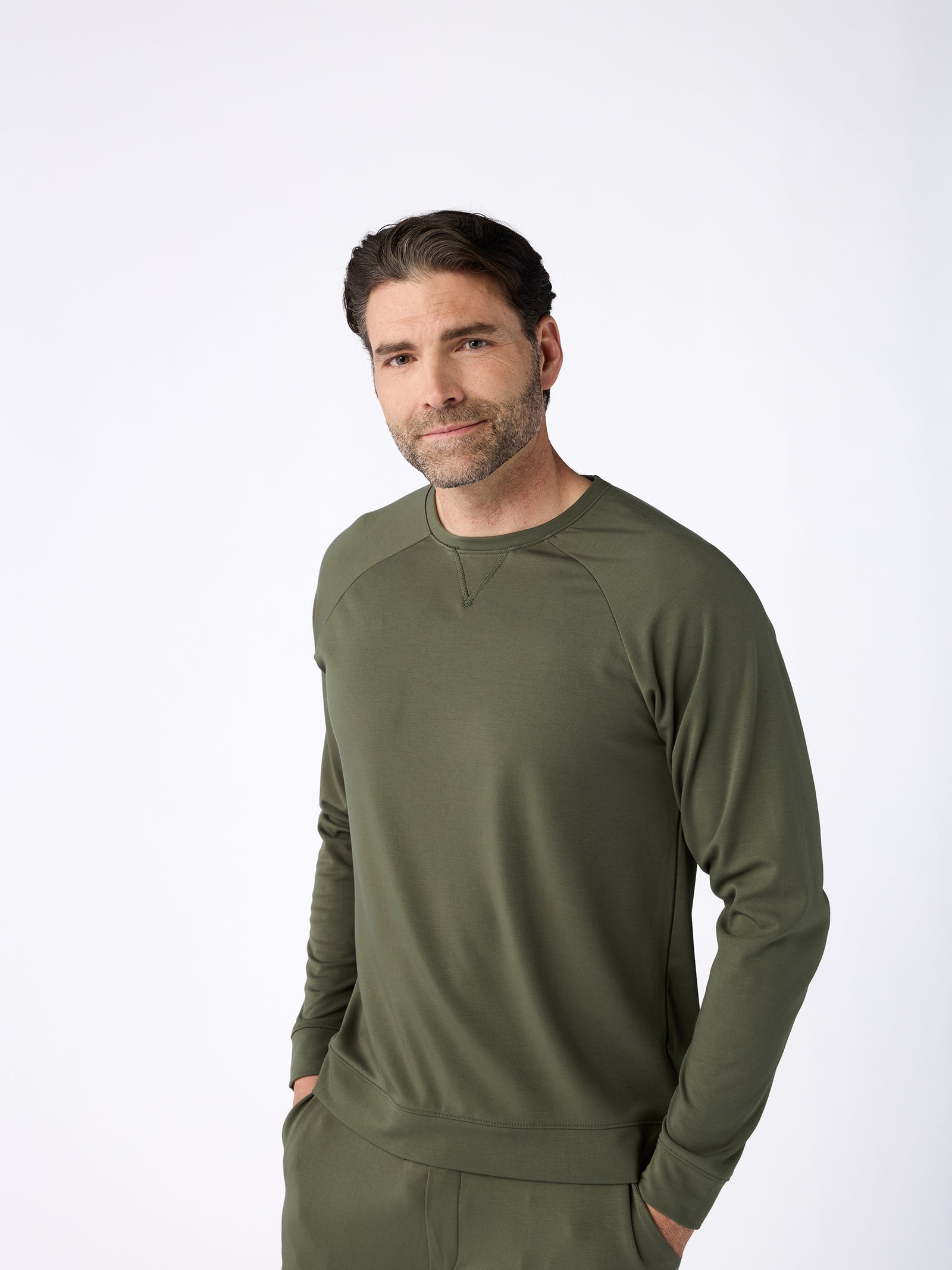 A man with dark hair and a beard is wearing the Men's Ultra-Soft Bamboo Pullover Crew from Cozy Earth in an olive green color. He is standing against a plain white background with his hands in his pockets, smiling slightly at the camera. 