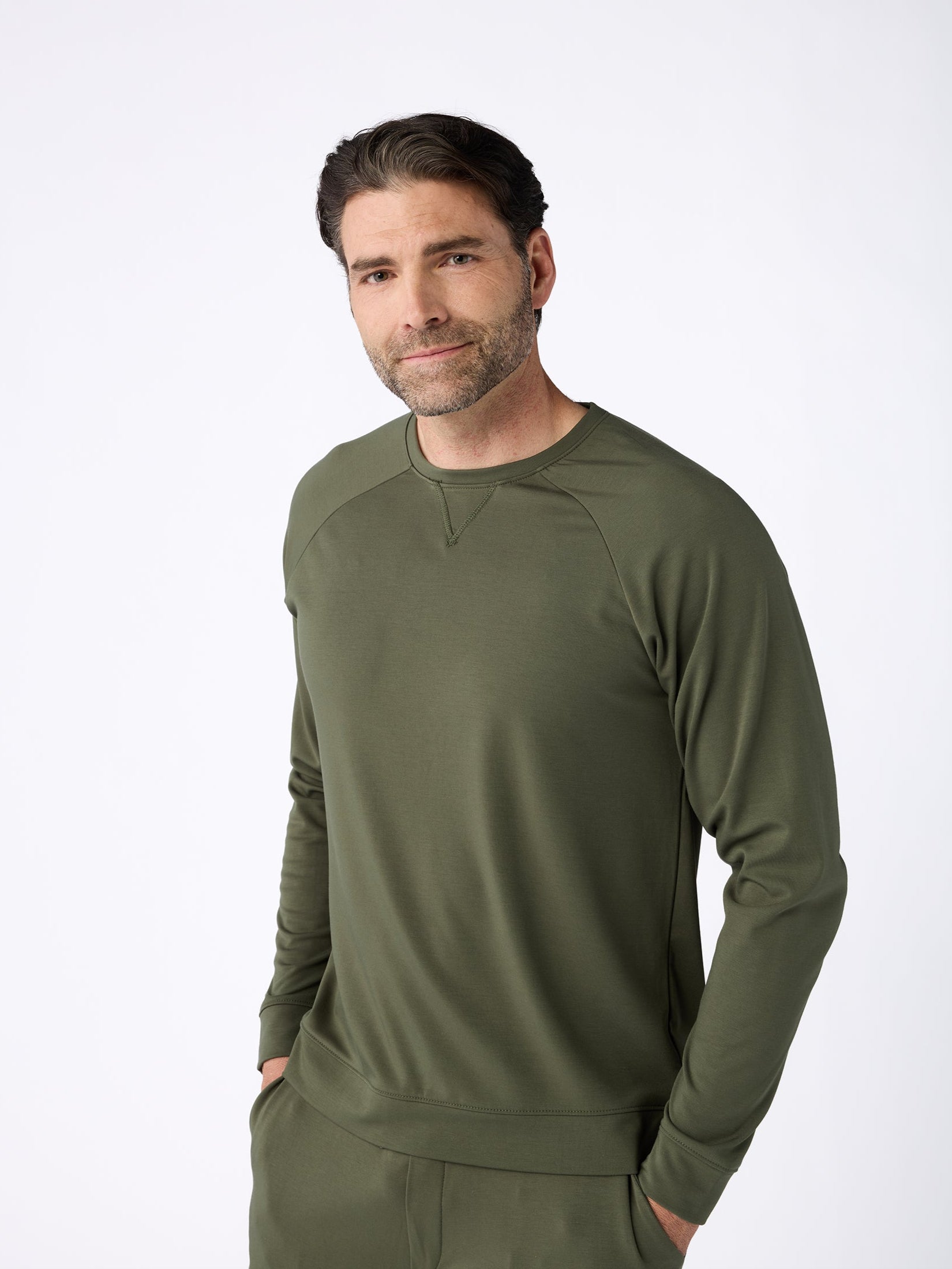 A man with dark hair and a beard is wearing the Men's Ultra-Soft Bamboo Pullover Crew from Cozy Earth in an olive green color. He is standing against a plain white background with his hands in his pockets, smiling slightly at the camera. |Color:Olive