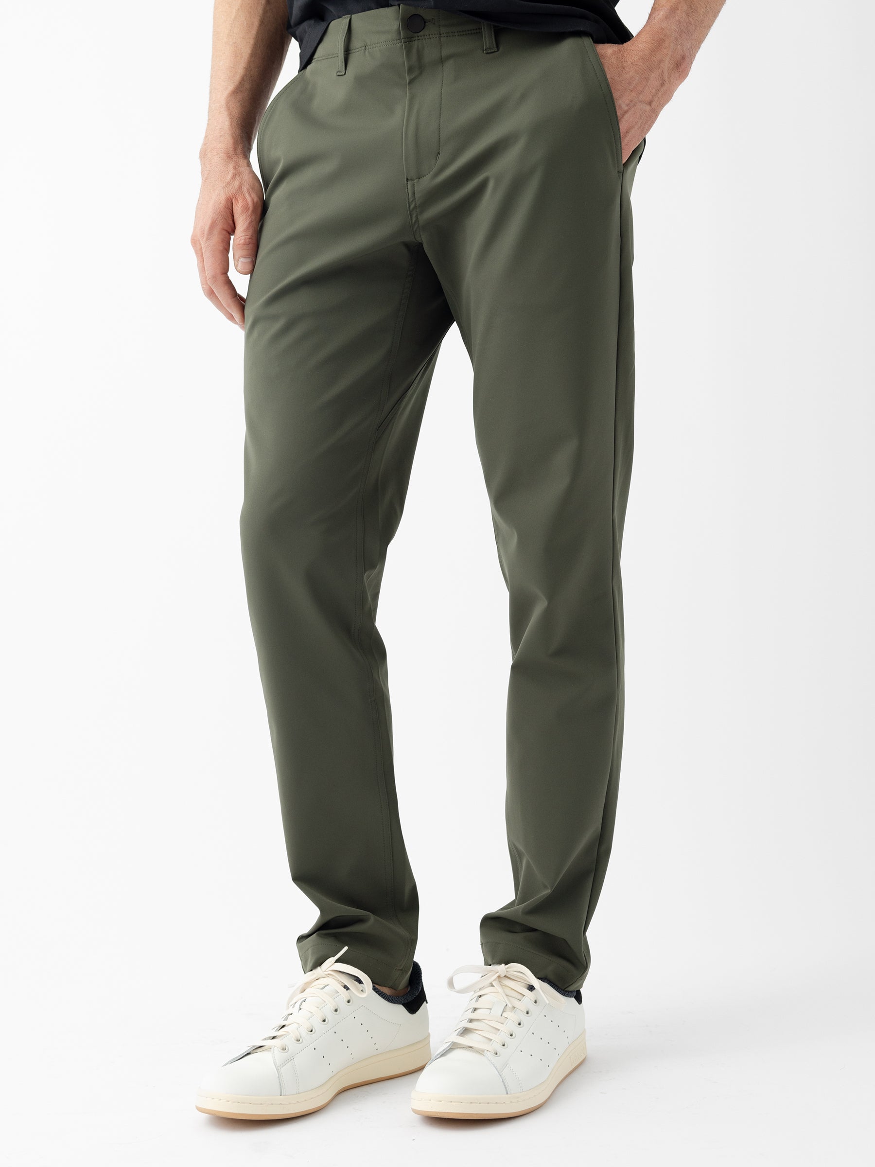 A person is shown wearing the Men's Everywhere Pant 32L from Cozy Earth in green along with white sneakers featuring black details. The background is plain white, and only the lower half of the body is visible. The individual's left hand rests by their side. |Color:Olive