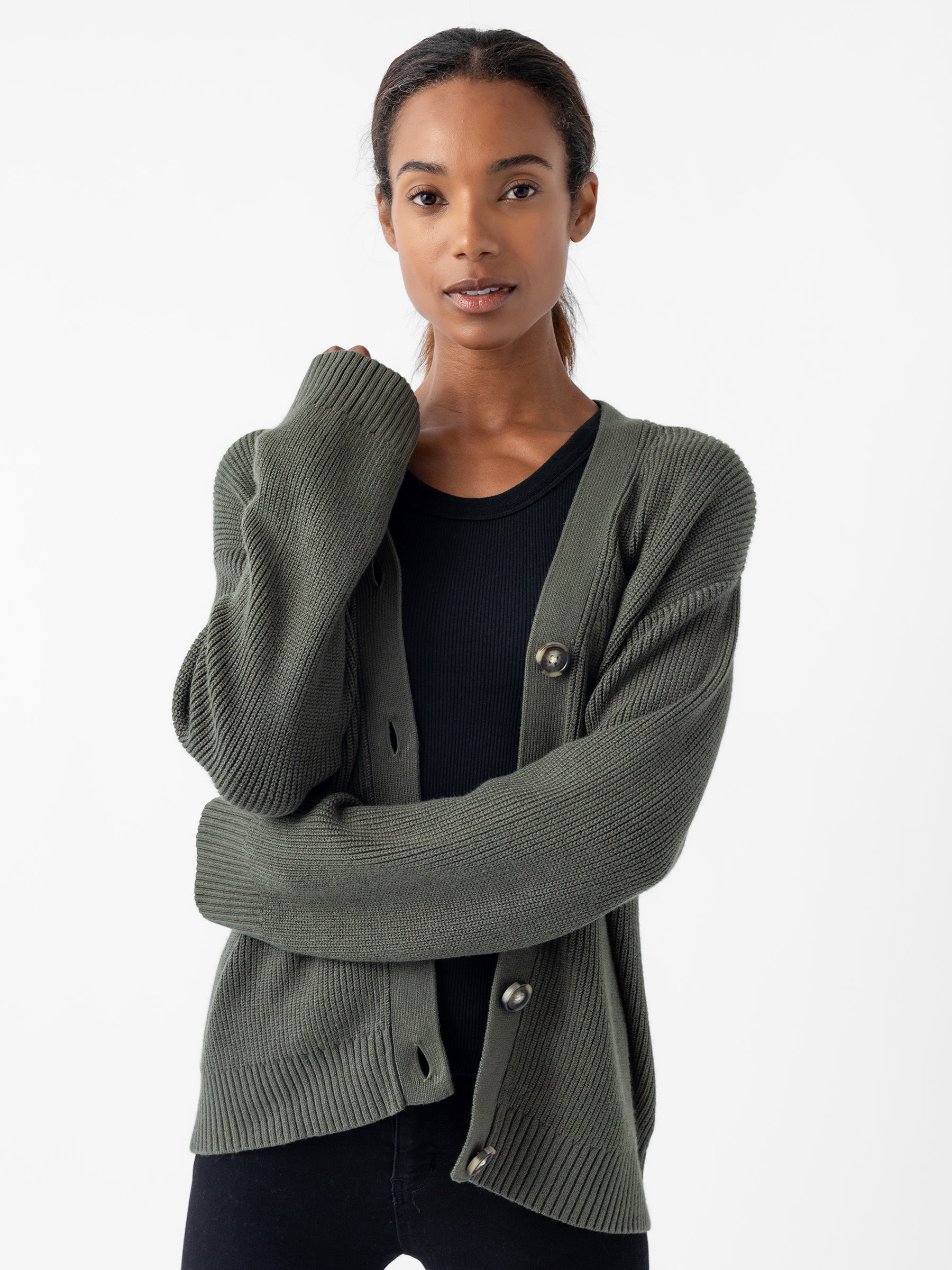 A person wearing a Cozy Earth Oversized Classic Cardigan in dark olive over a black shirt, with long dark hair pulled back. They are standing with their arms slightly crossed against a plain white background. |Color:Olive