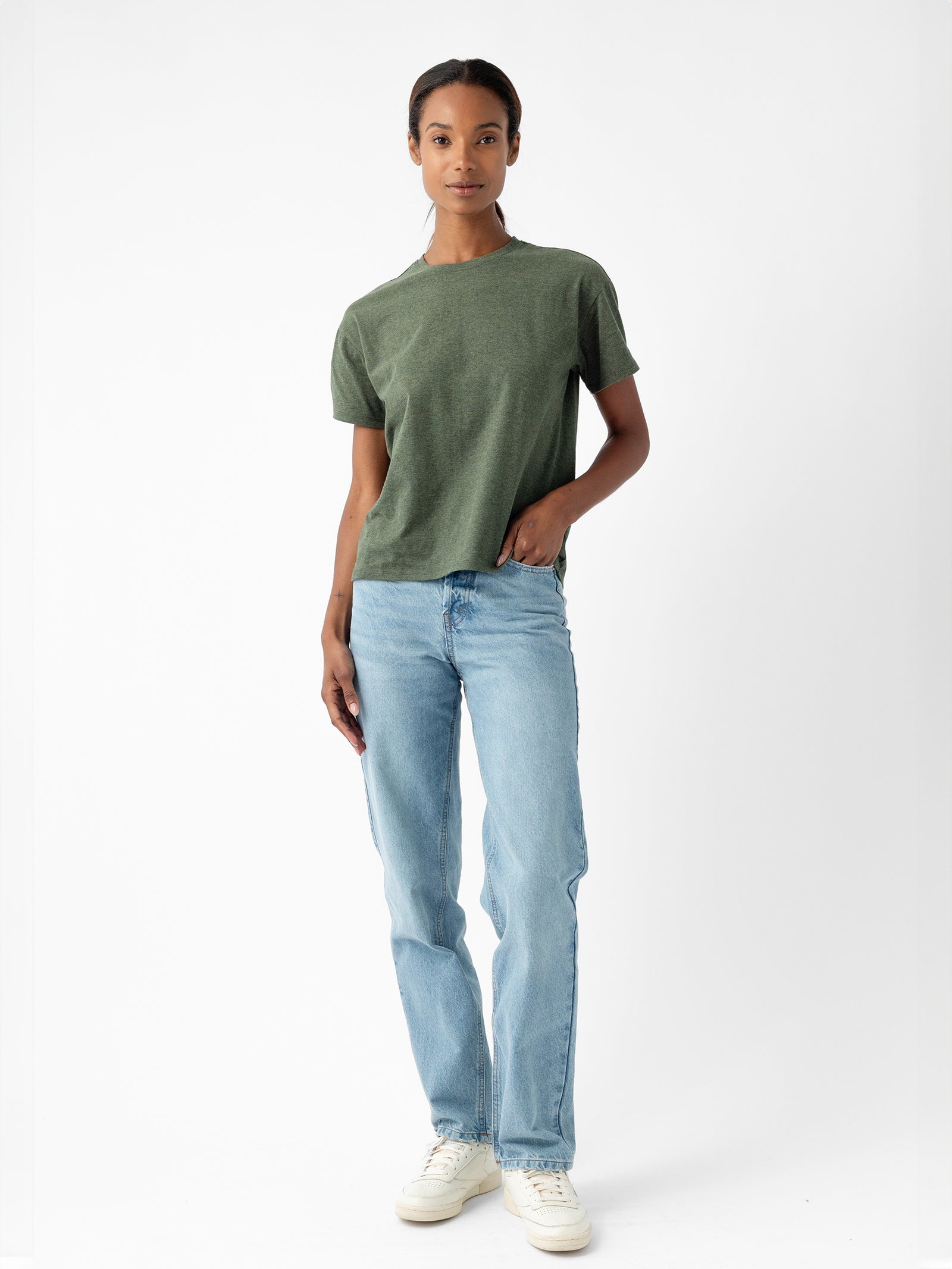 Against a plain white background, a person dressed in Cozy Earth's Women's All Day Tee, which is short-sleeved and green, pairs it with light blue jeans and white sneakers. One hand hangs relaxed by their side while the other rests casually in their front pocket. Their expression is calm and neutral. 