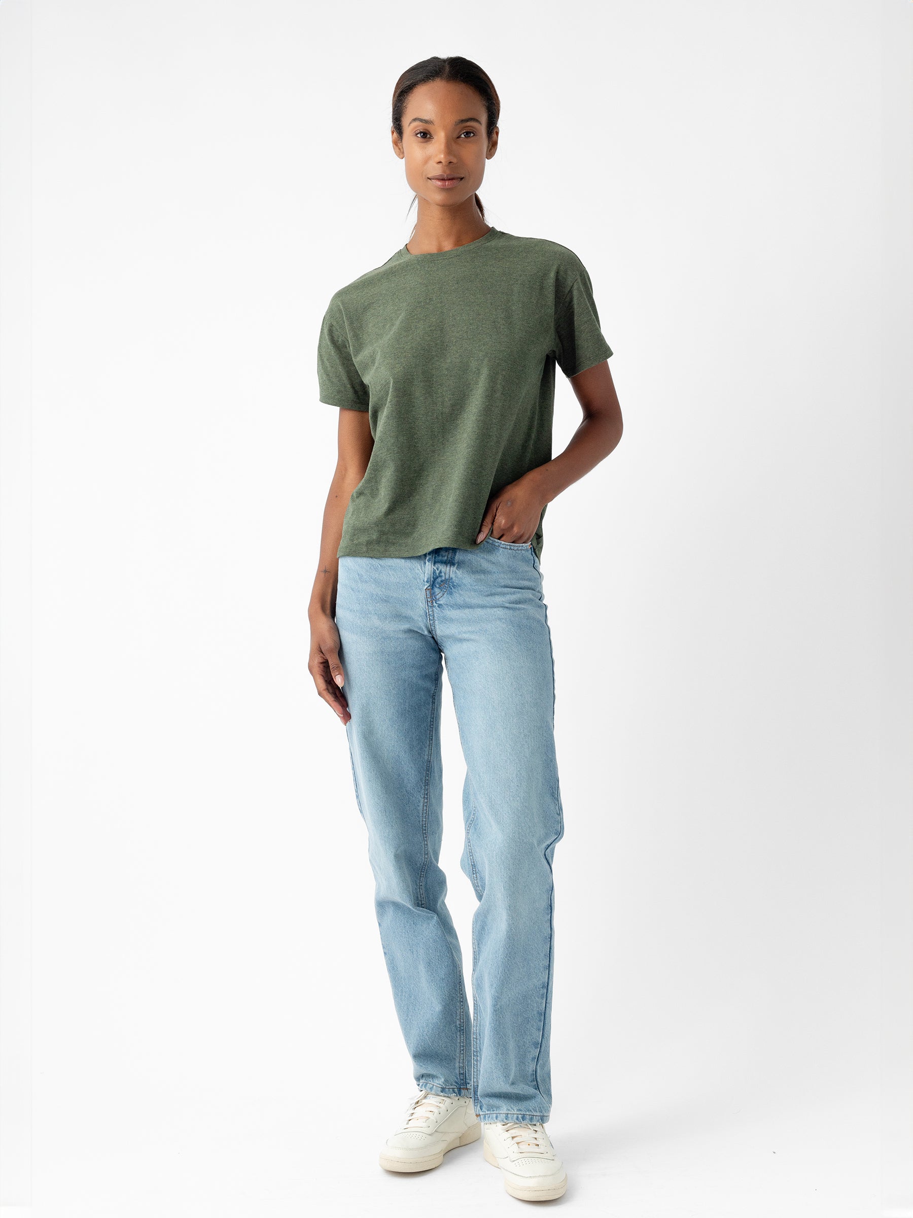 Against a plain white background, a person dressed in Cozy Earth's Women's All Day Tee, which is short-sleeved and green, pairs it with light blue jeans and white sneakers. One hand hangs relaxed by their side while the other rests casually in their front pocket. Their expression is calm and neutral. |Color:Olive Heather