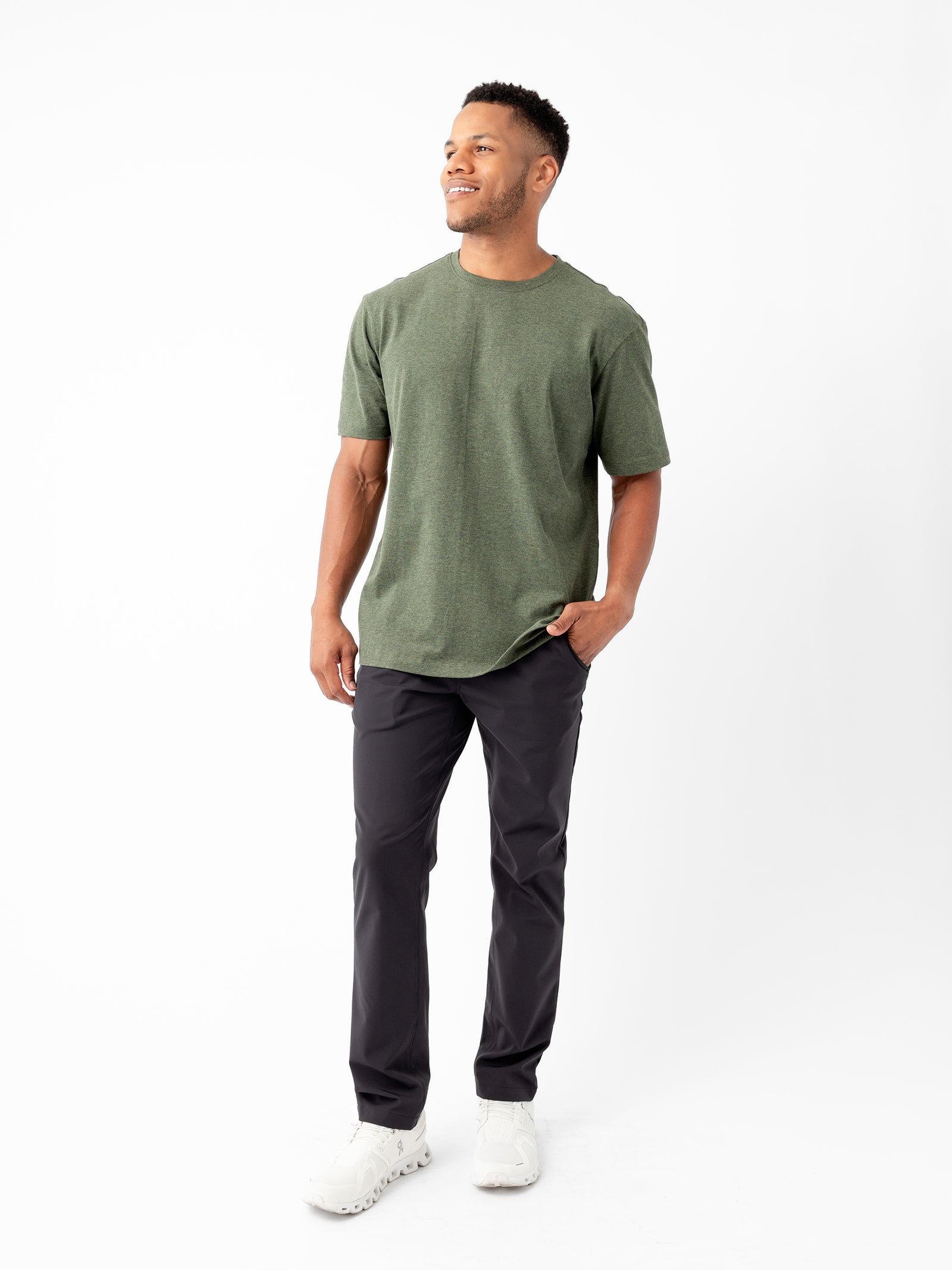 A man is smiling while standing against a plain white background, wearing a Cozy Earth Men's All Day Tee in green, paired with black pants and white sneakers. One of his hands is in his pocket while the other rests relaxed at his side. 