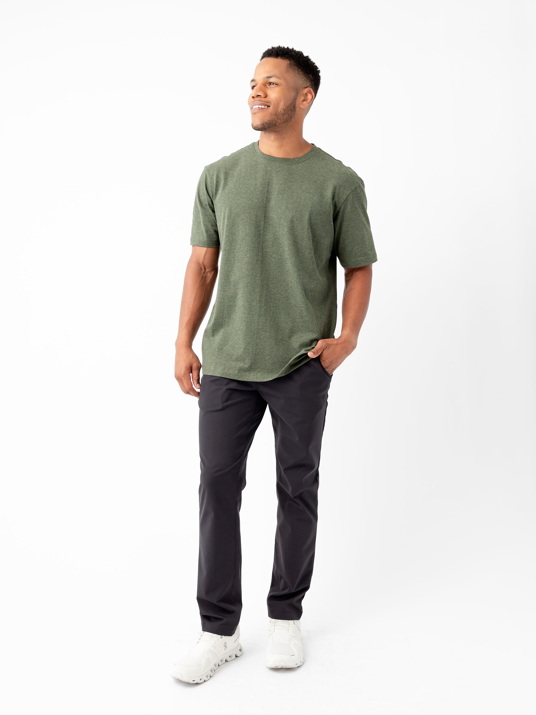 A man is smiling while standing against a plain white background, wearing a Cozy Earth Men's All Day Tee in green, paired with black pants and white sneakers. One of his hands is in his pocket while the other rests relaxed at his side. |Color:Olive Heather