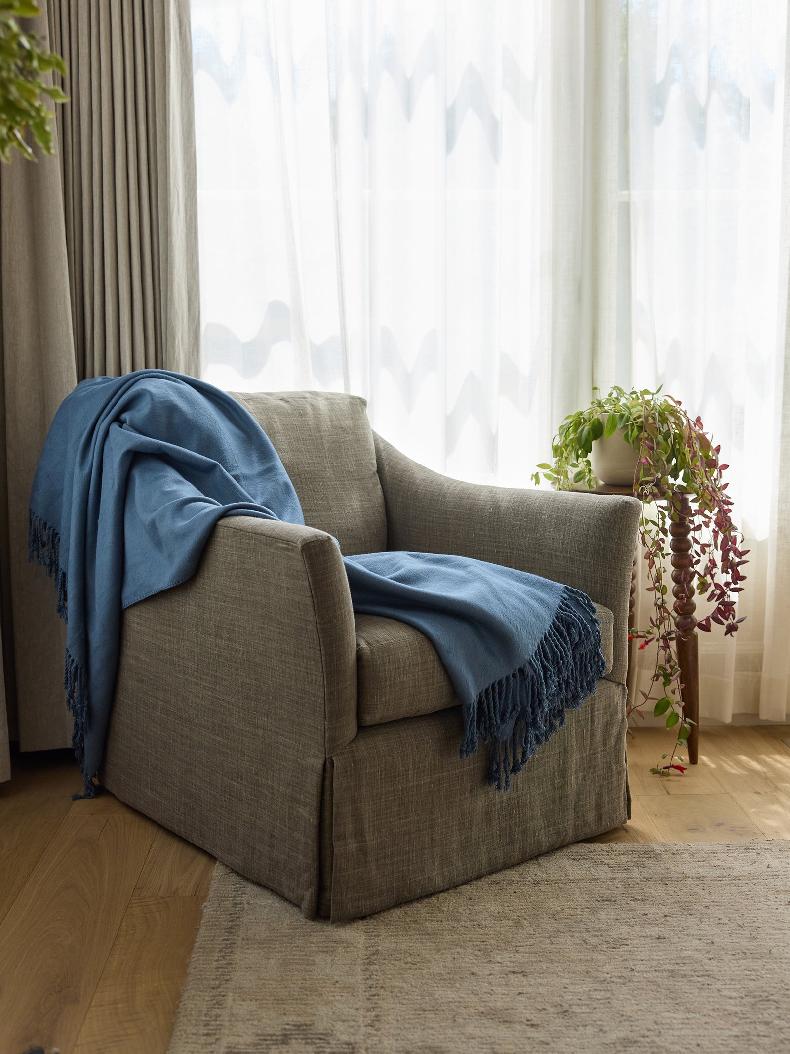 A cozy gray armchair draped with a Cozy Earth Bamboo Tassel Throw sits by a large window with sheer curtains. Next to the chair is a potted plant with trailing vines on a stand. The setup is on a wooden floor with a light-colored rug nearby. 