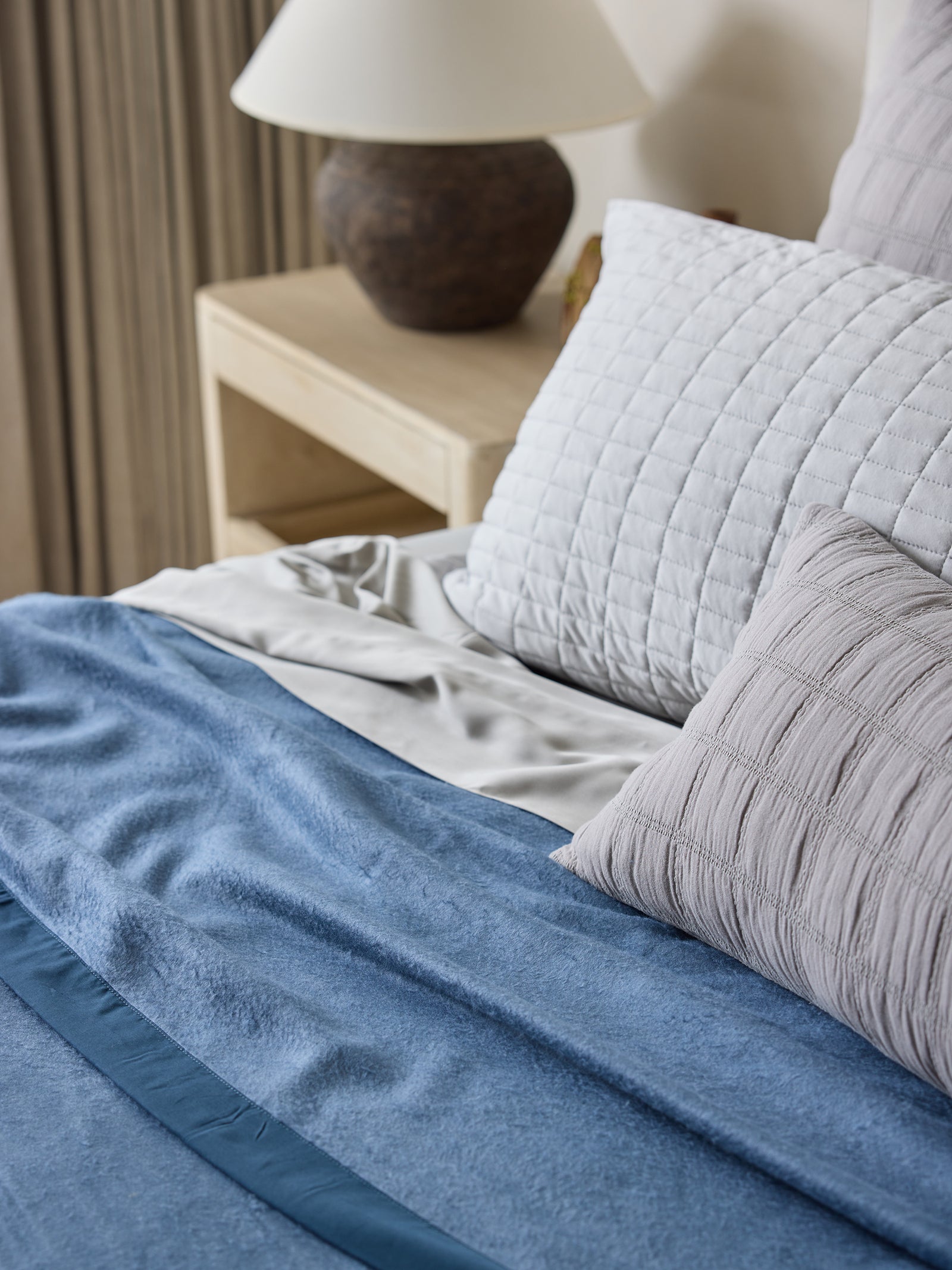 A neatly made bed featuring a Cozy Earth Bamboo Blanket in blue, gray textured pillows, and white sheets. A small, light-colored wooden nightstand with a ceramic lamp and a decorative item sits beside the bed. The background includes elegant, neutral-colored curtains. 