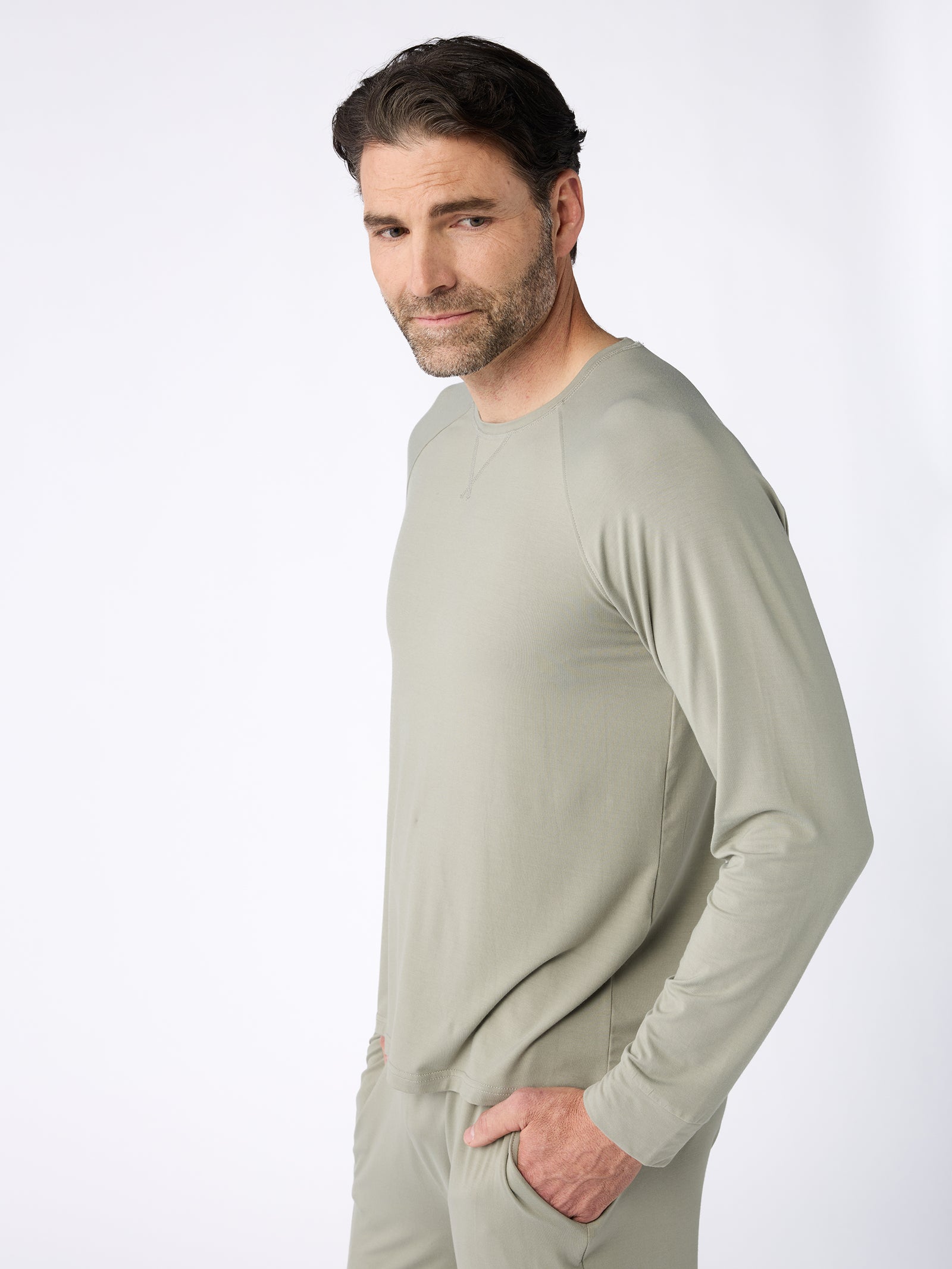 A man with short dark hair and a beard is dressed in a light gray Men's Stretch-Knit Bamboo Long Sleeve by Cozy Earth, paired with matching pants. He stands sideways with one hand in his pocket, glancing over his shoulder against a plain white background. 