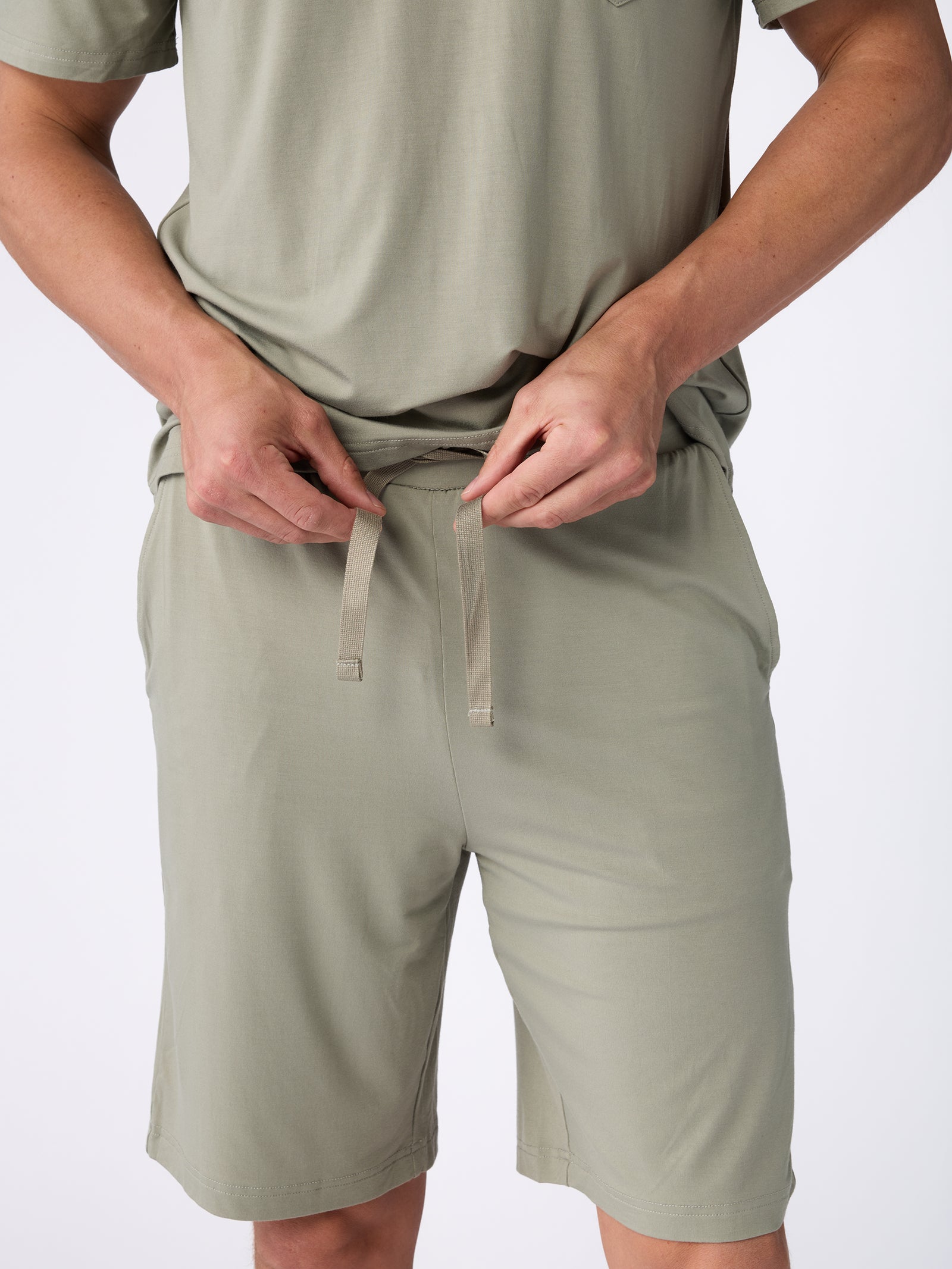 Wearing a light green ensemble, an individual adjusts the drawstring of their **Men's Stretch-Knit Bamboo Pajama Short** from **Cozy Earth**. Both the shorts and T-shirt are crafted from a smooth fabric, set against a plain white background. 