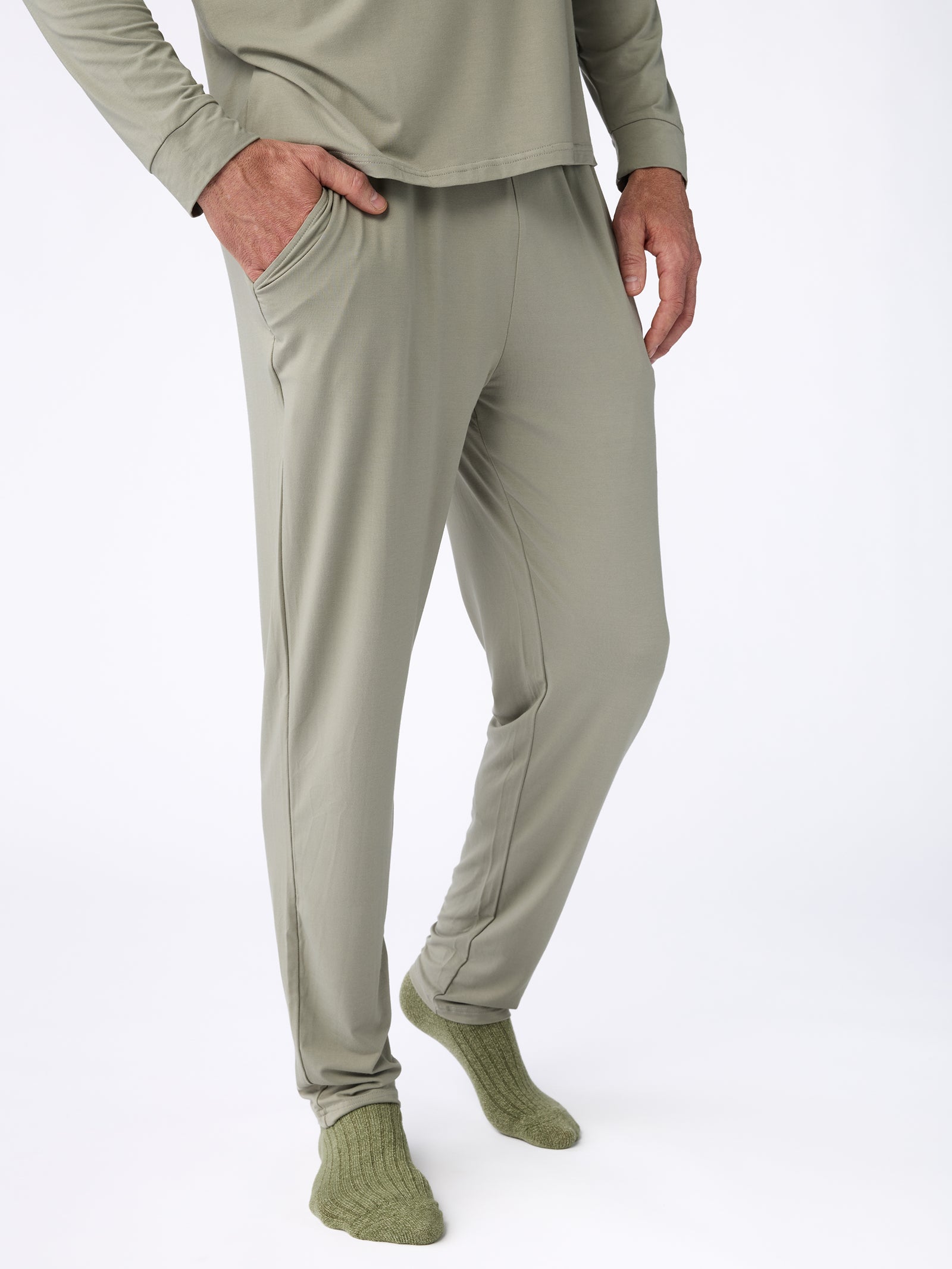 A person is dressed in Cozy Earth's Men's Stretch-Knit Bamboo Pajama Pant in a light green color, complete with long pants and a coordinating long-sleeve top. With one hand resting casually in their pants pocket, they complement the outfit with green socks against a plain white background. 