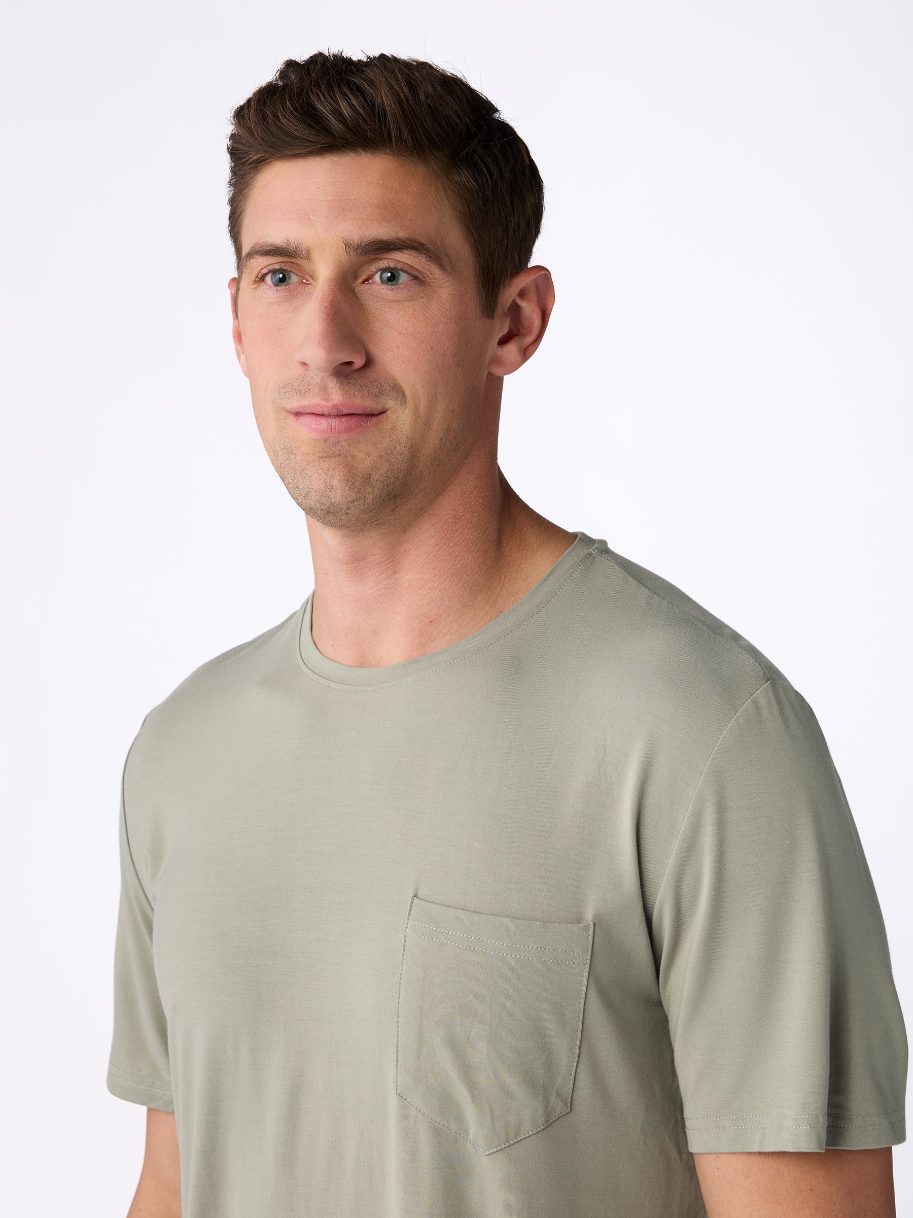 A man wearing the Men's Stretch-Knit Bamboo Lounge Tee by Cozy Earth, in a light gray color featuring a chest pocket, stands against a plain white background. He gazes slightly to the side, with short brown hair and a neutral expression. |Color:Palm