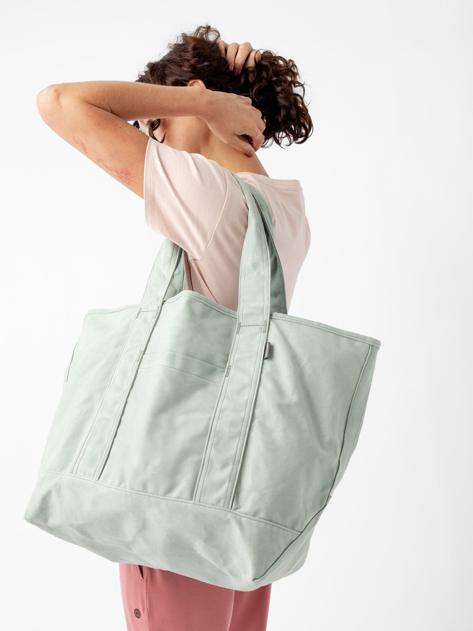 A person with short, curly hair is scratching their head while holding a large, light green Waxed Canvas Tote from Cozy Earth over their shoulder. They are wearing a light pink T-shirt and pink pants, standing against a plain white background. 