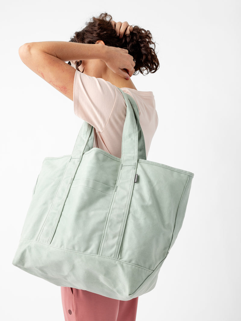 A person with short, curly hair is scratching their head while holding a large, light green Waxed Canvas Tote from Cozy Earth over their shoulder. They are wearing a light pink T-shirt and pink pants, standing against a plain white background.