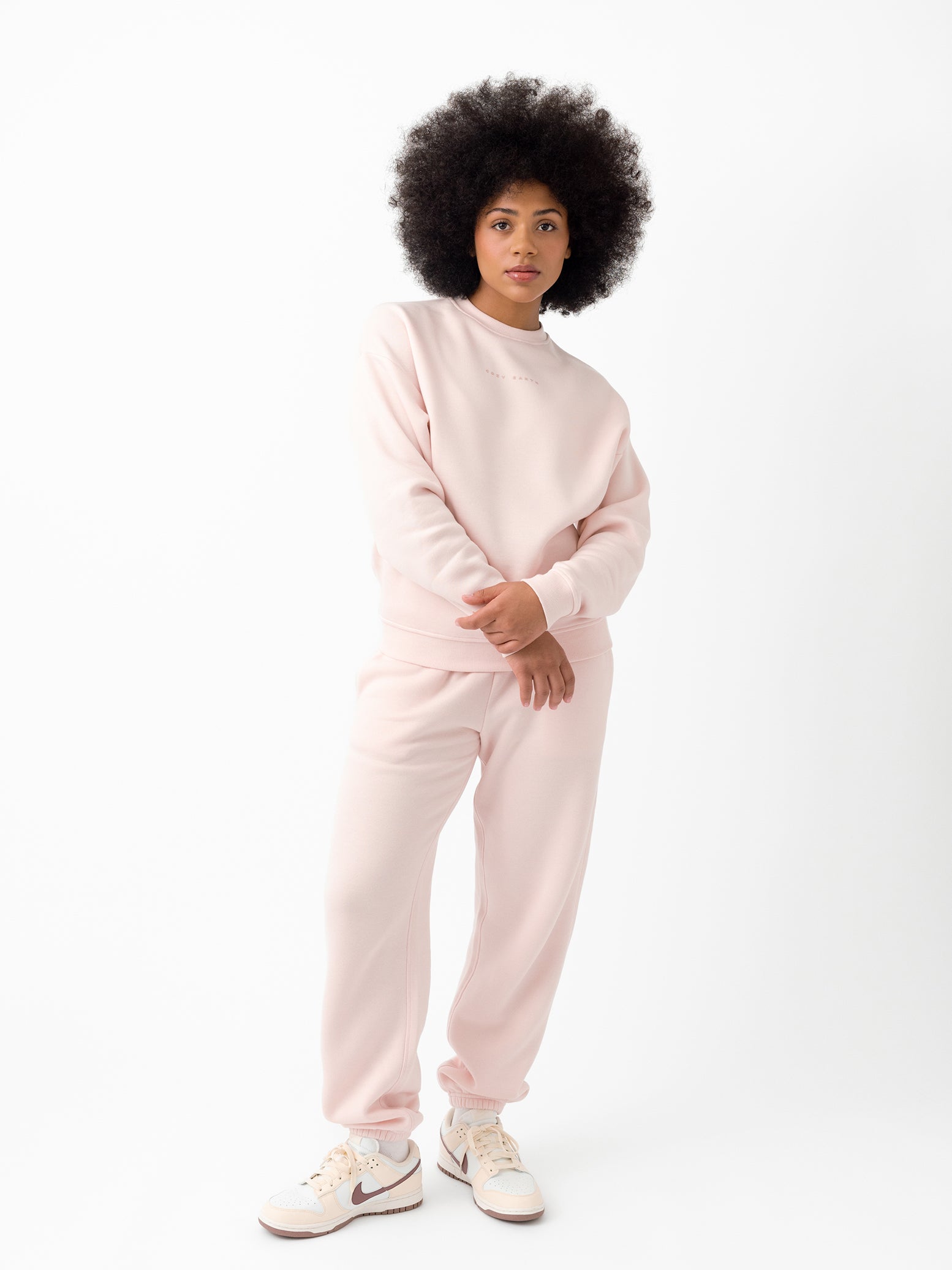 Dressed in Cozy Earth's Women's CityScape Crewneck and matching sweatpants, a person with curly hair stands against a plain white backdrop. They're sporting white sneakers with a purple swoosh while gazing toward the camera. 