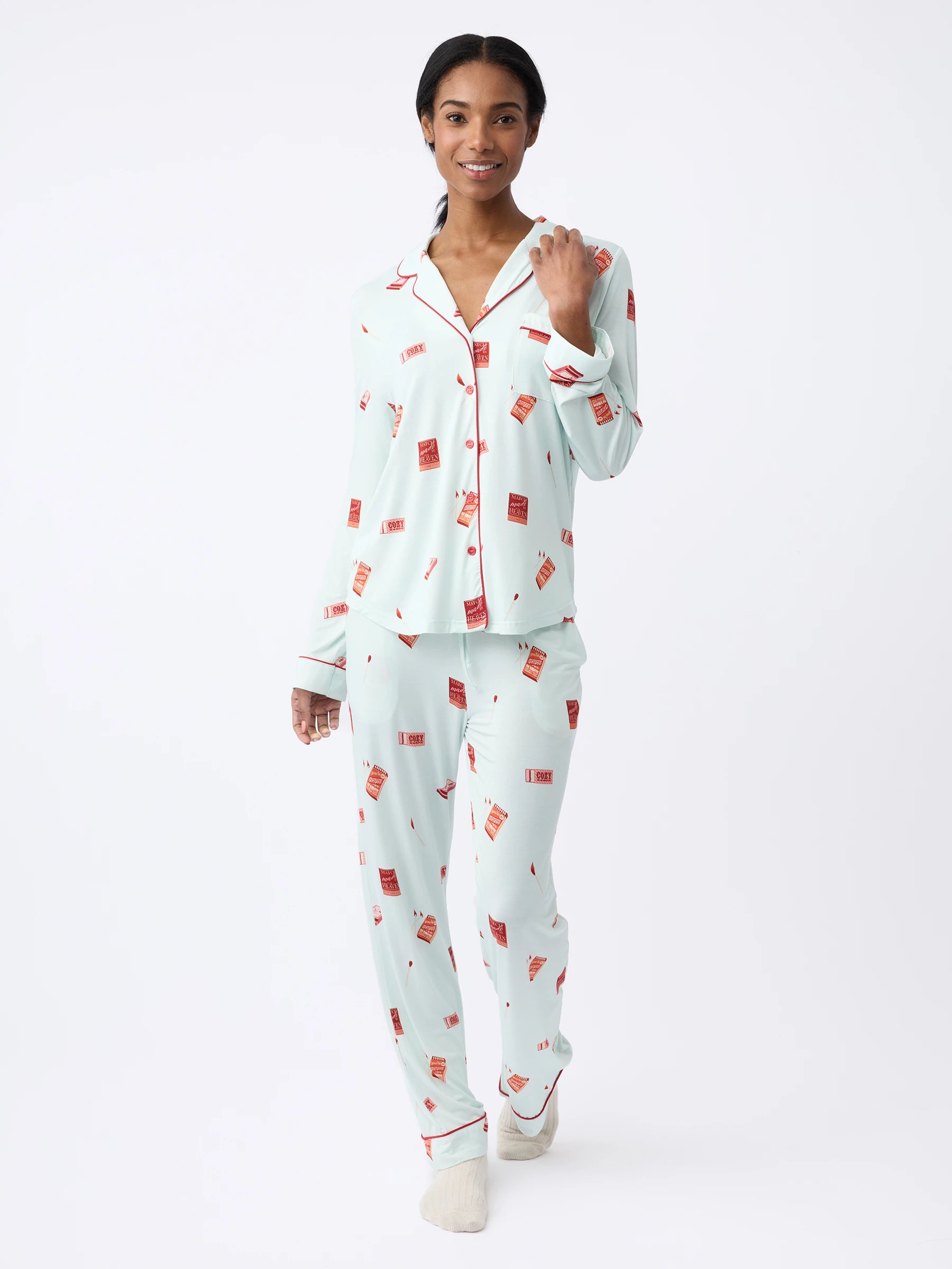 A woman poses on a white background in Cozy Earth's Women's Stretch-Knit Long Sleeve Bamboo Pajama Set, featuring light blue fabric with red and white patterns. The set includes a button-up top and matching pants, and the person looks relaxed and cheerful. |Color:Perfect Match