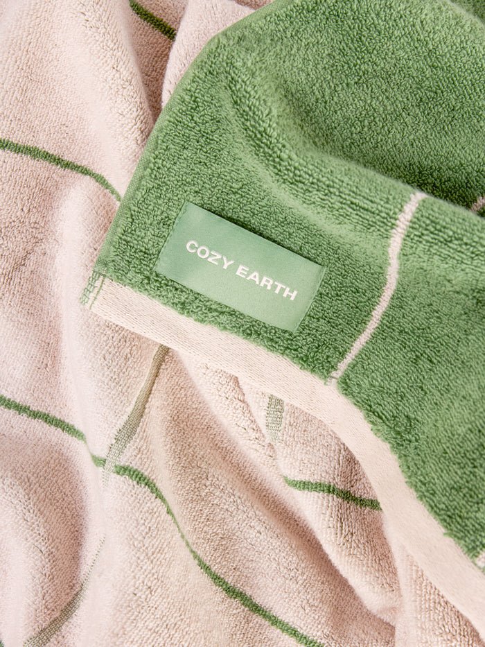 A close-up of the Windowpane Resort Towel by Cozy Earth shows a green and beige design with thin stripes. The soft, plush towel features a neatly stitched edge and a "COZY EARTH" label. 