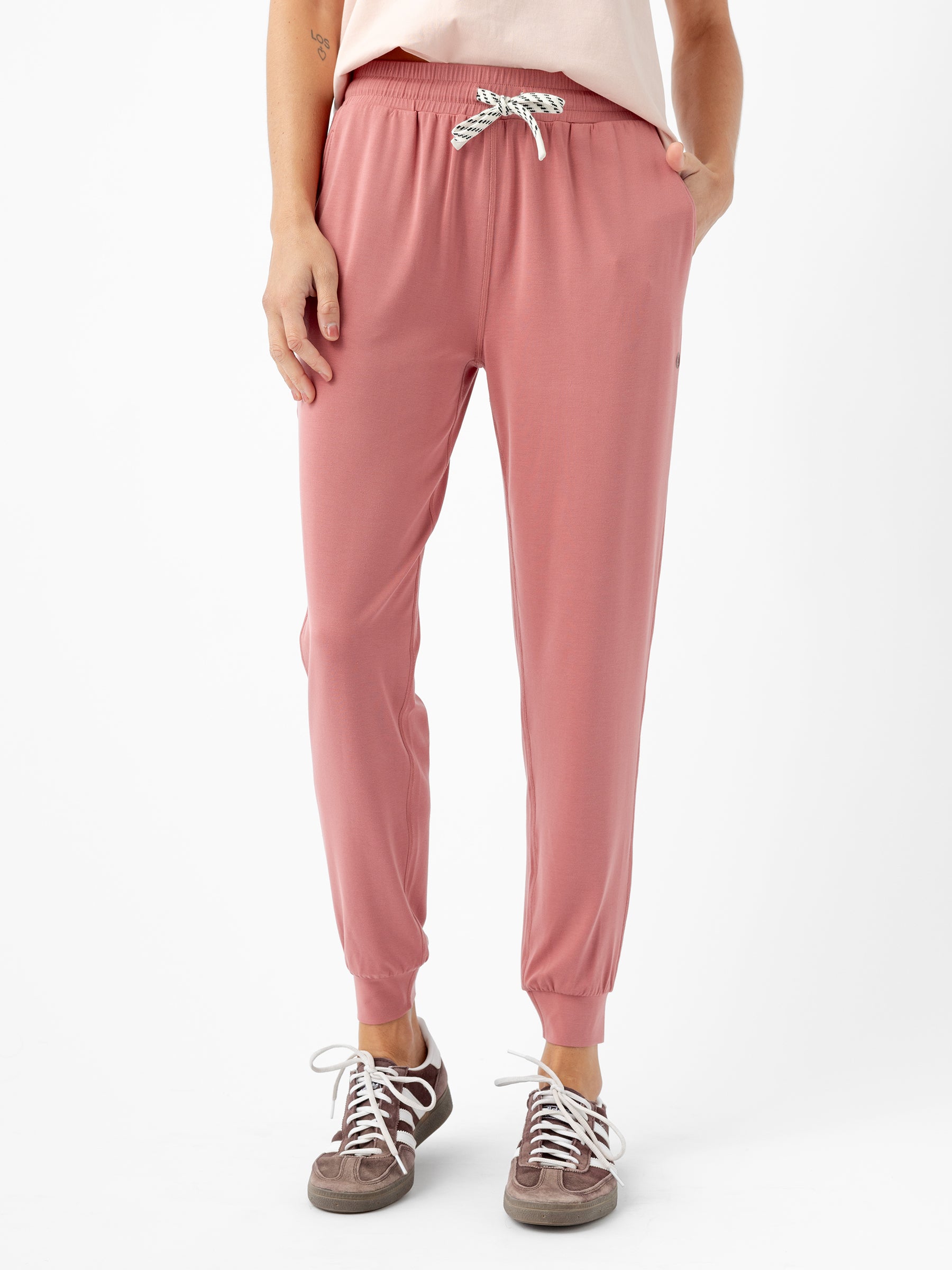 A person is standing and wearing Cozy Earth's Women's Studio Jogger, which features a pink color with a white drawstring and pockets where their hands rest. They are also dressed in a light-colored top and grey sneakers with white laces against a white background. 