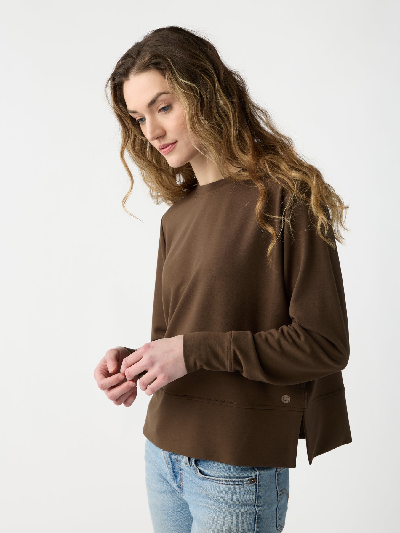 A person with long wavy hair is wearing Cozy Earth's Women's StudioLite Crewneck and blue jeans, standing against a plain background, looking slightly downward with a calm expression. |Color:Sable