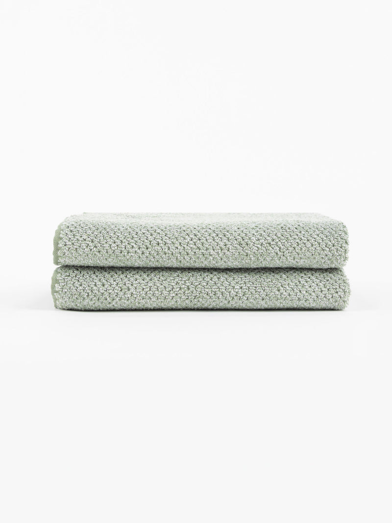 Two Nantucket Bath Towels by Cozy Earth, in a light green textured finish, are neatly folded and stacked on a white surface with a plain white background.