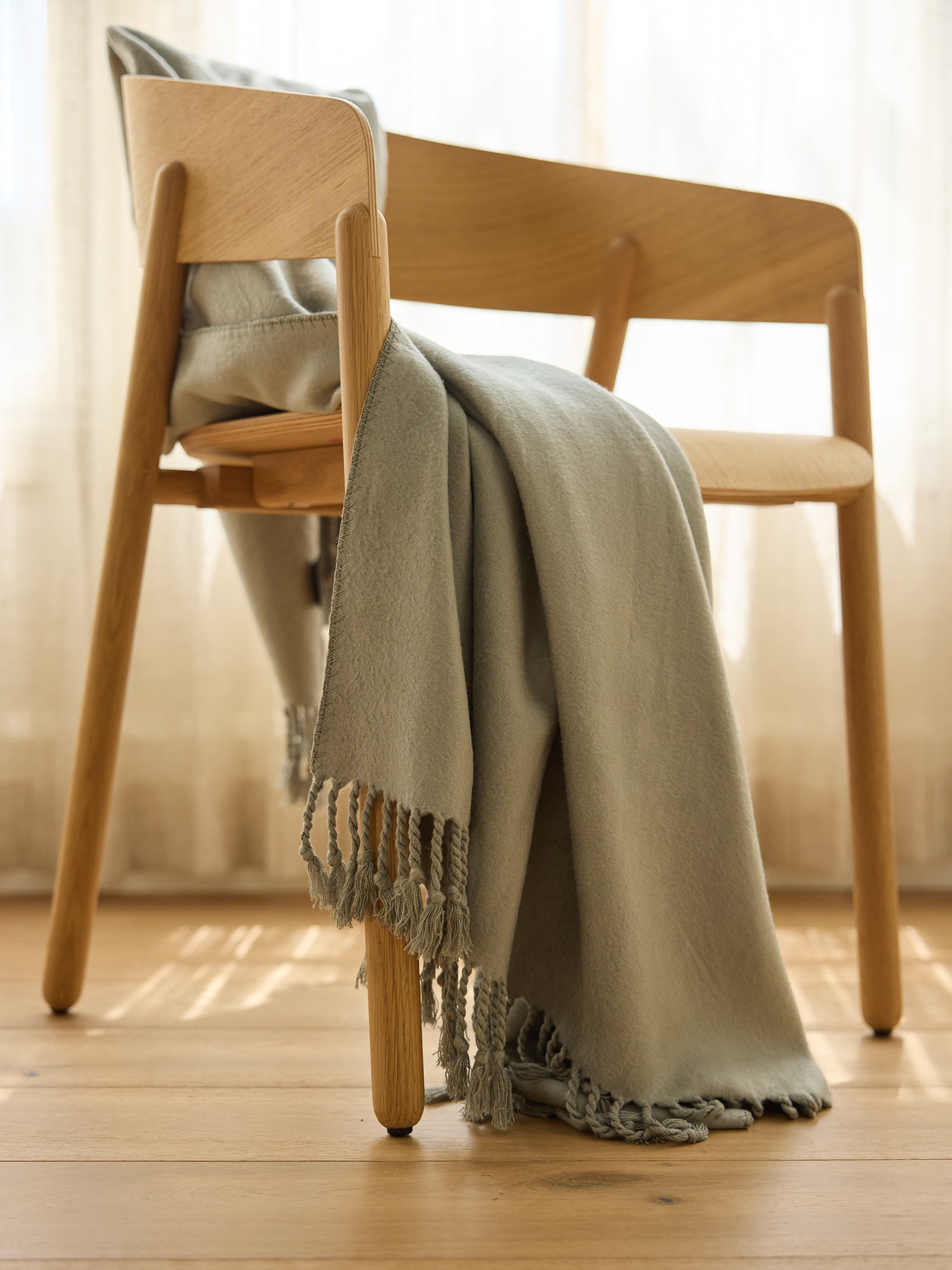 A light wooden chair with a minimalist design is partially covered by the Bamboo Tassel Throw from Cozy Earth. The scene is set in a warmly lit room with light-colored curtains and wooden flooring, creating a cozy and inviting atmosphere. 