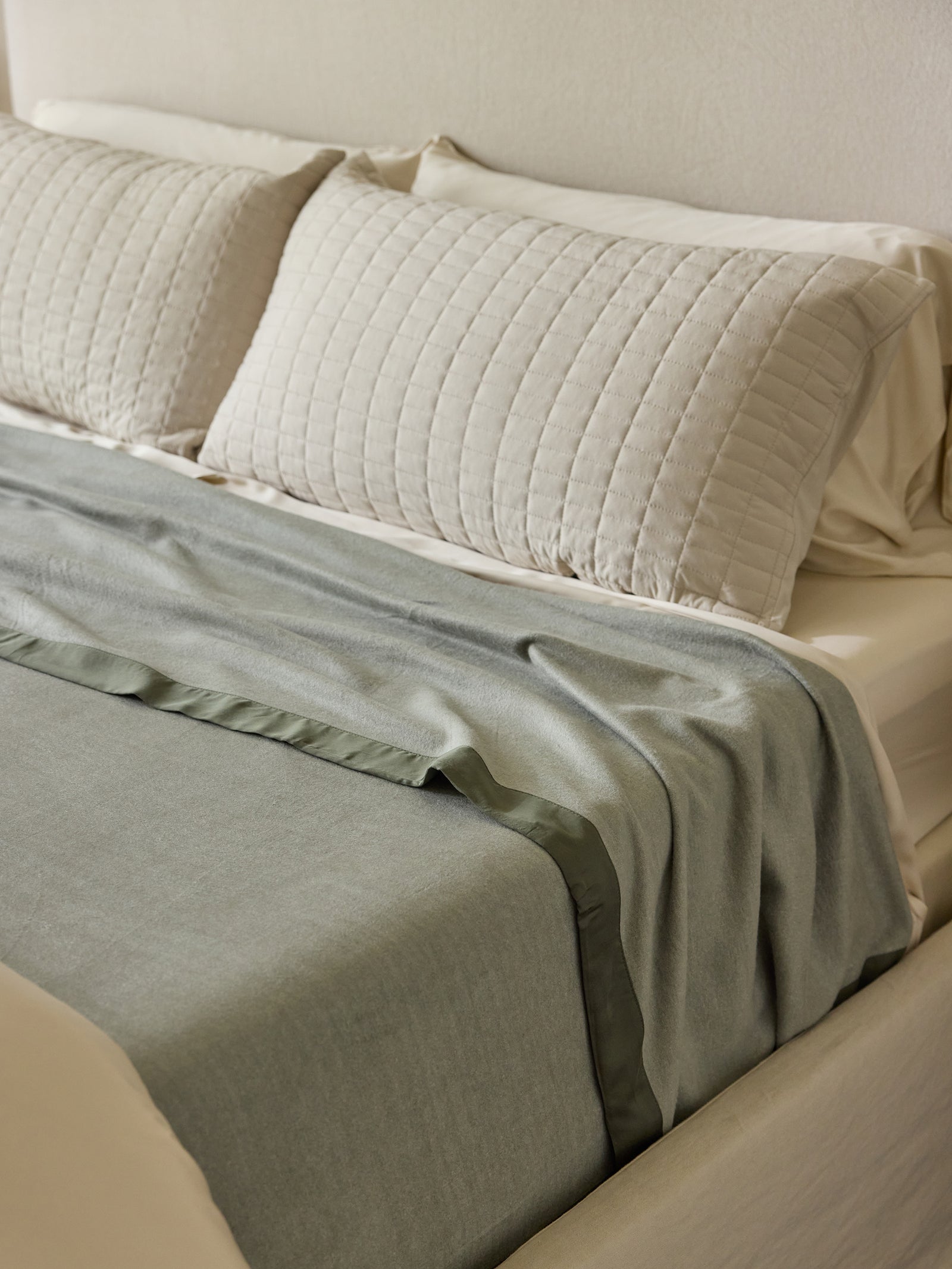 A neatly made bed features two white quilted pillows against a beige headboard, covered with light gray sheets and a matching gray duvet with a dark green hem, folded back slightly to reveal a white mattress. The overall look is clean and minimalistic, complemented by the luxurious feel of the Bamboo Blanket from Cozy Earth. 