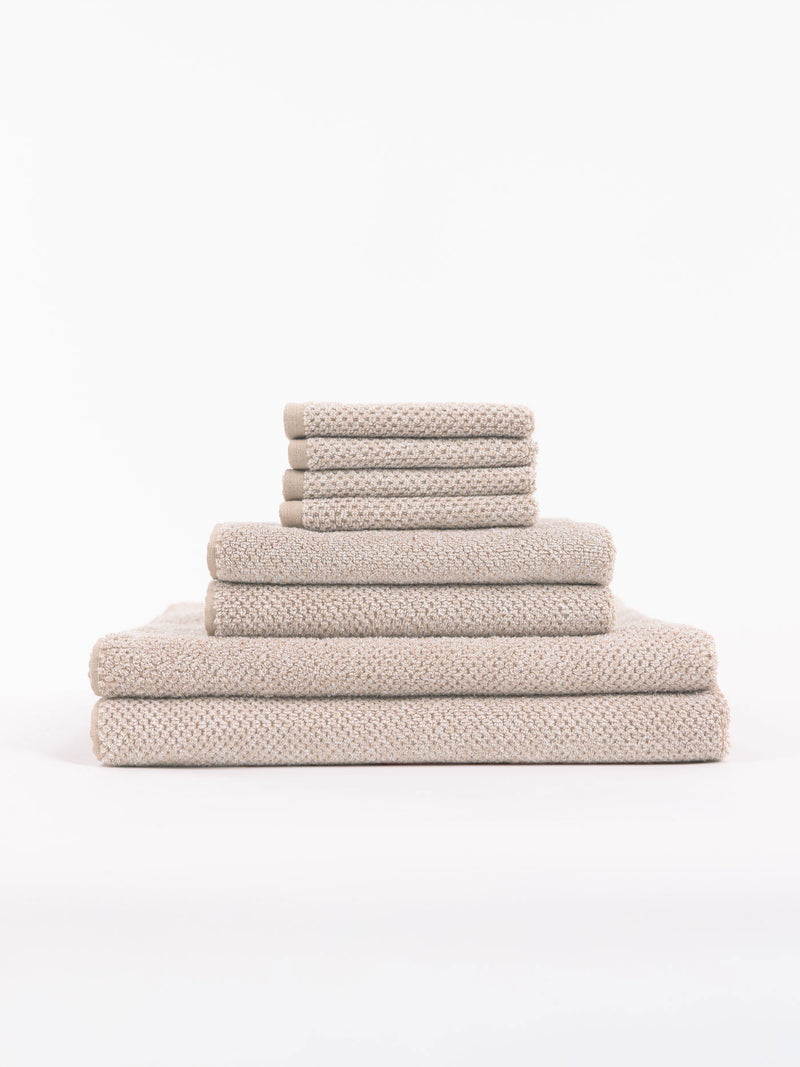 The Cozy Earth Nantucket Bath Towel Set features beige textured towels in various sizes, neatly stacked from largest to smallest, set against a plain white background.