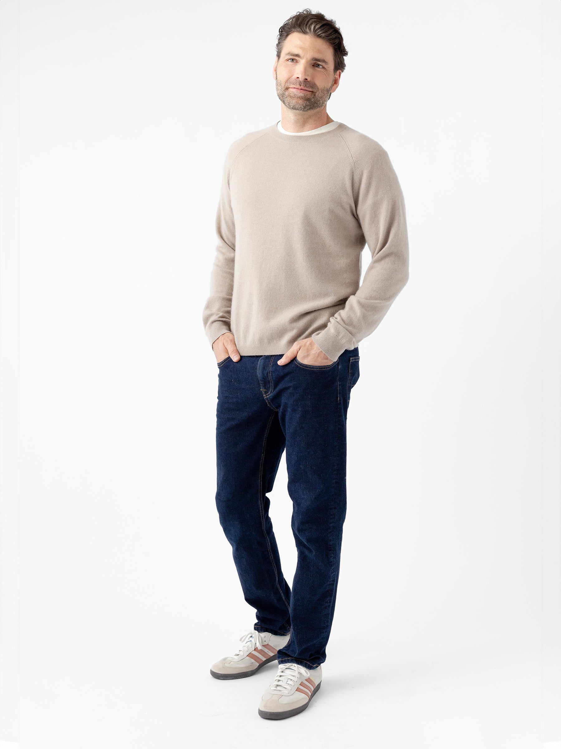 A man stands against a white background wearing a Men's Crewneck Sweater by Cozy Earth, dark blue jeans, and gray sneakers with white and red accents. He has his hands in his pockets and is smiling slightly at the camera. |Color:Sandstone