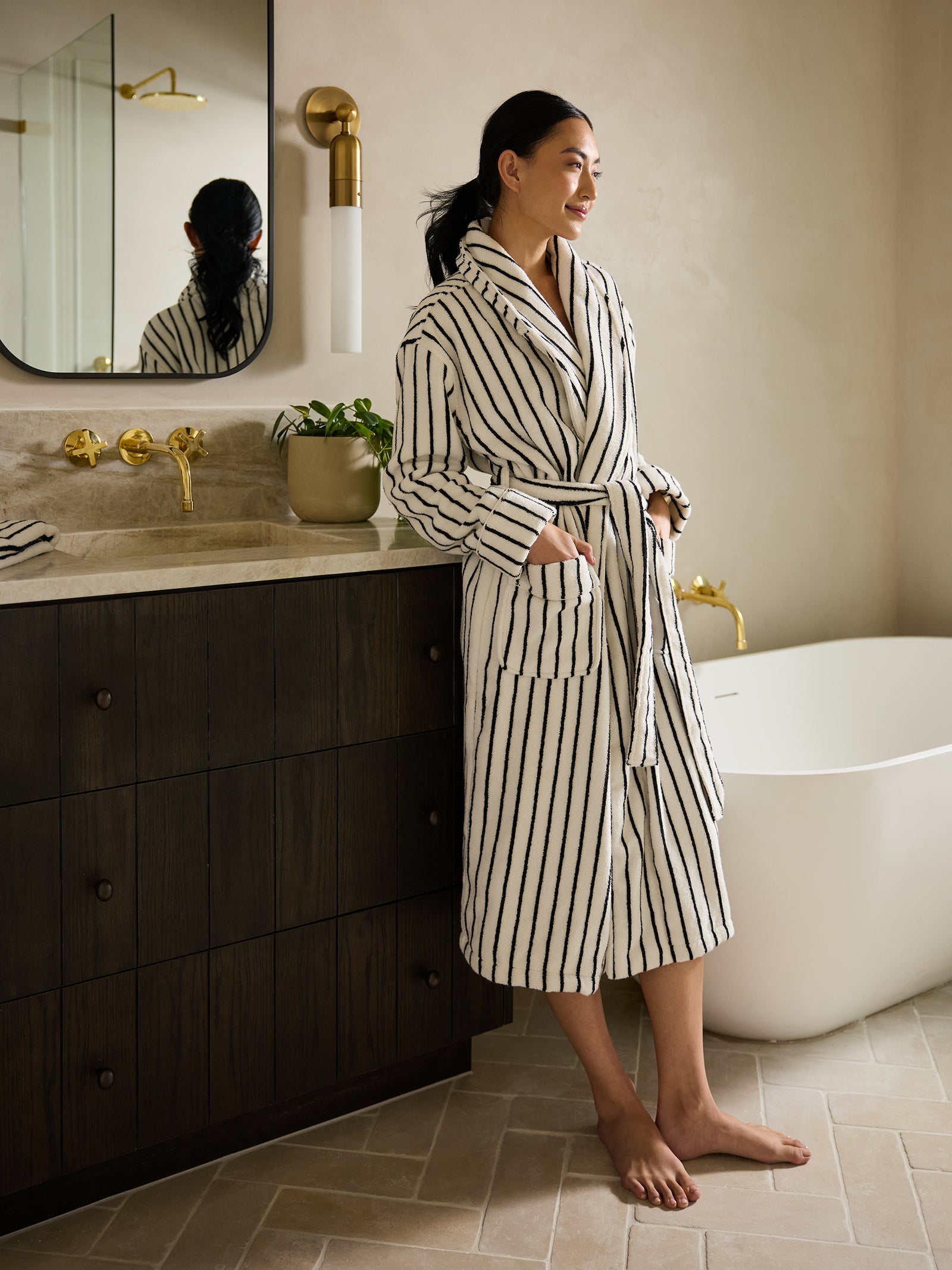 A person wearing a Luxe Bath Robe by Cozy Earth stands in a modern bathroom, leaning against a dark wood vanity. Nearby, there is a white bathtub and a potted plant, with the bathroom also featuring a large mirror and gold fixtures. 