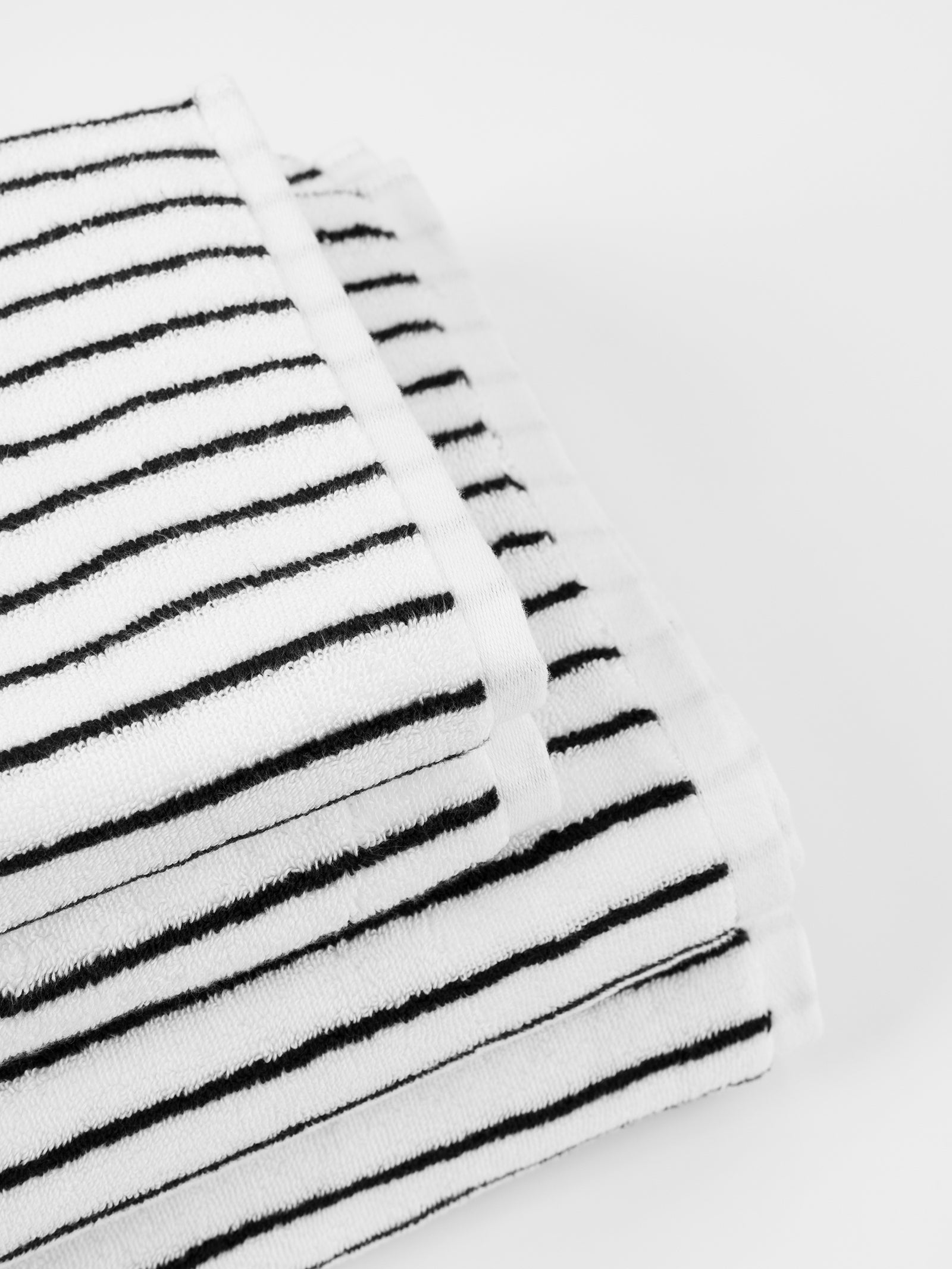 A close-up view of Luxe Bath Towels by Cozy Earth, neatly folded with thin black stripes, placed on a plain white surface. The towels feature a soft texture and a clean, modern design pattern. 