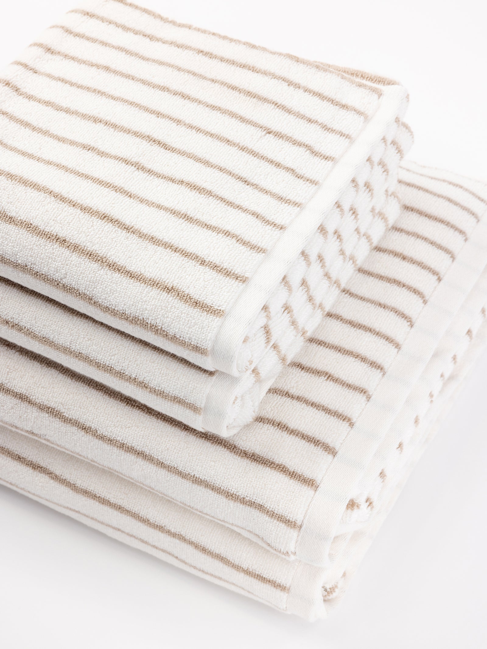 A stack of three Luxe Bath Towels by Cozy Earth in white with light brown stripes are neatly arranged, with each towel slightly offset from the one beneath it. The simple, elegant design gives a clean and inviting appearance. 