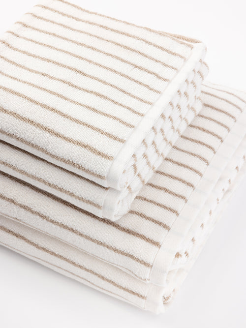 A stack of neatly folded Luxe Hand Towels from Cozy Earth, featuring light brown horizontal stripes on a white background. The towels are arranged in descending size order, with the largest at the bottom and the smallest at the top. The scene is set against a clean, white surface. |Color:Sand Stripe