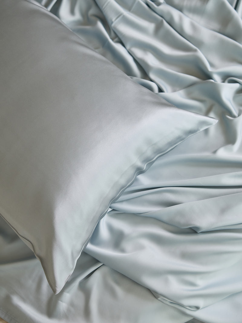 A smooth, pale blue Silk Pillowcase from Cozy Earth rests on a matching satin sheet, with the fabric softly gathered around the pillow, creating a luxurious and tranquil appearance.