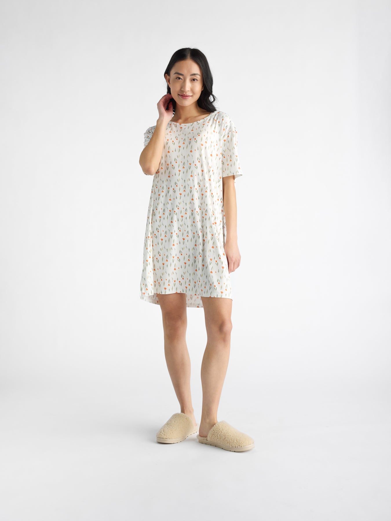 A person wearing Cozy Earth's Women's Bamboo Stretch-Knit Sleep Dress and beige fluffy slippers smiles, touching their hair against a plain white background. |Color:Sienna Tulips