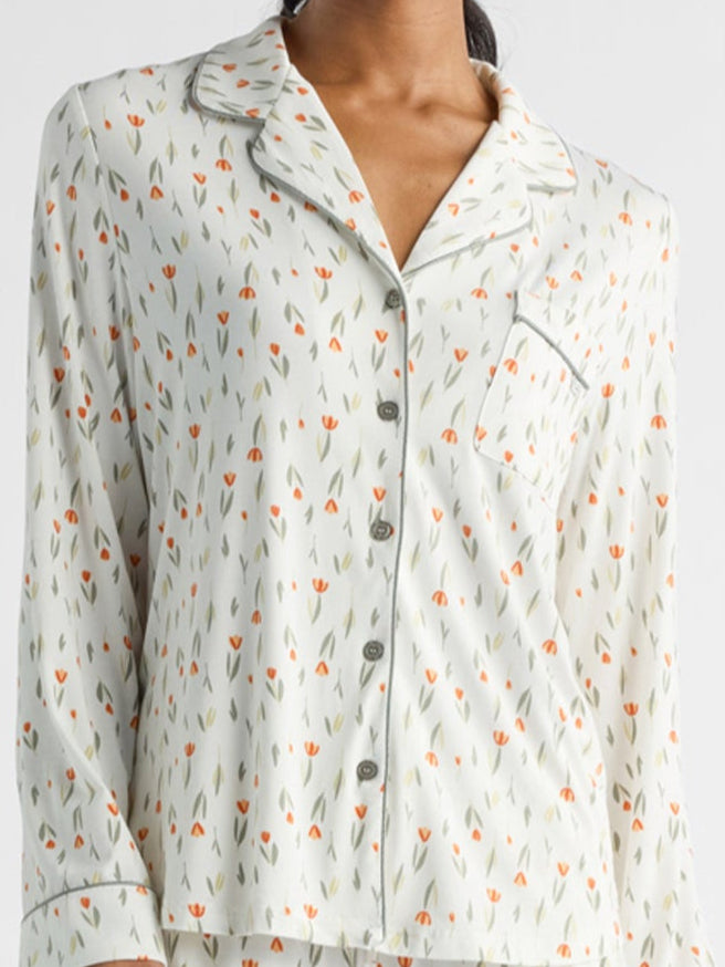 A person wearing a pajama top adorned with a small floral pattern featuring orange and green tulips. The top has a button-up front, a collar, and breast pocket. The background is plain white.