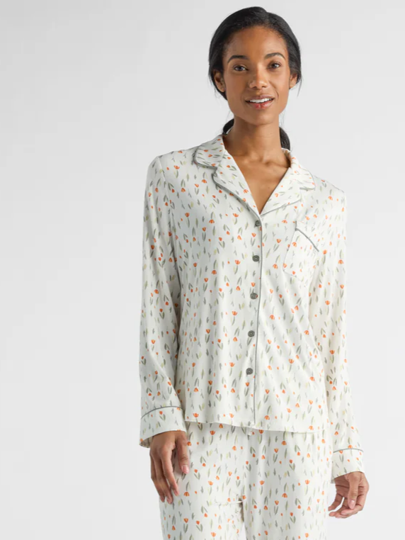A person is wearing a matching pajama set with a pattern of small, colorful abstract shapes on a light background. They are standing and smiling with hands by their sides, against a plain, neutral backdrop.