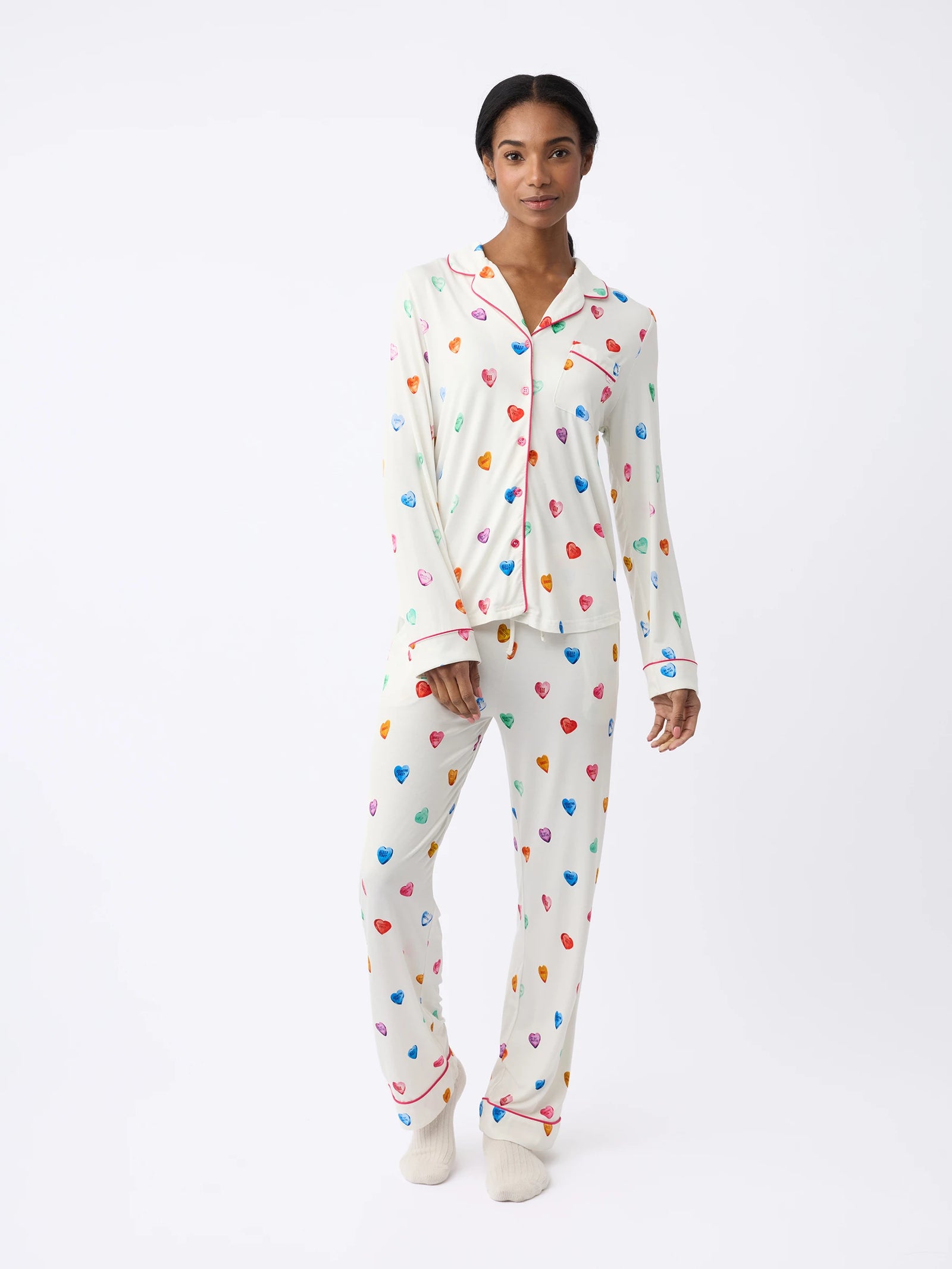 A person in a Women's Long Sleeve Bamboo Pajama Top by HIDE in stretch-knit, featuring colorful heart patterns, stands against a plain white background. The set includes matching pants and cream-colored socks.