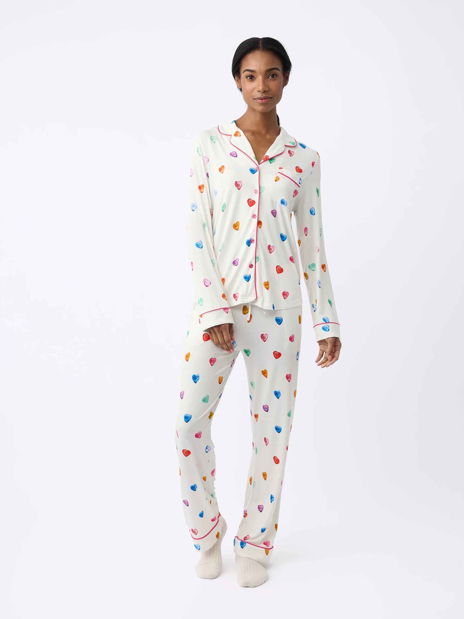 A person wears a white Women's Stretch-Knit Bamboo Pajama Pant by Cozy Earth with colorful heart patterns, featuring a button-up shirt with a collar and matching pants, against a plain background. They also wear light-colored socks. 