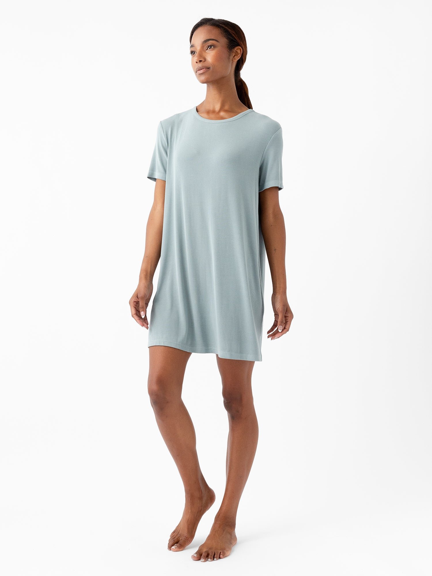 A woman stands barefoot against a white background, wearing the light blue, knee-length, short-sleeved Women’s Bamboo Rib-Knit Boyfriend Sleep Dress by Cozy Earth. She has long hair and appears relaxed, with her arms gently hanging by her sides. 